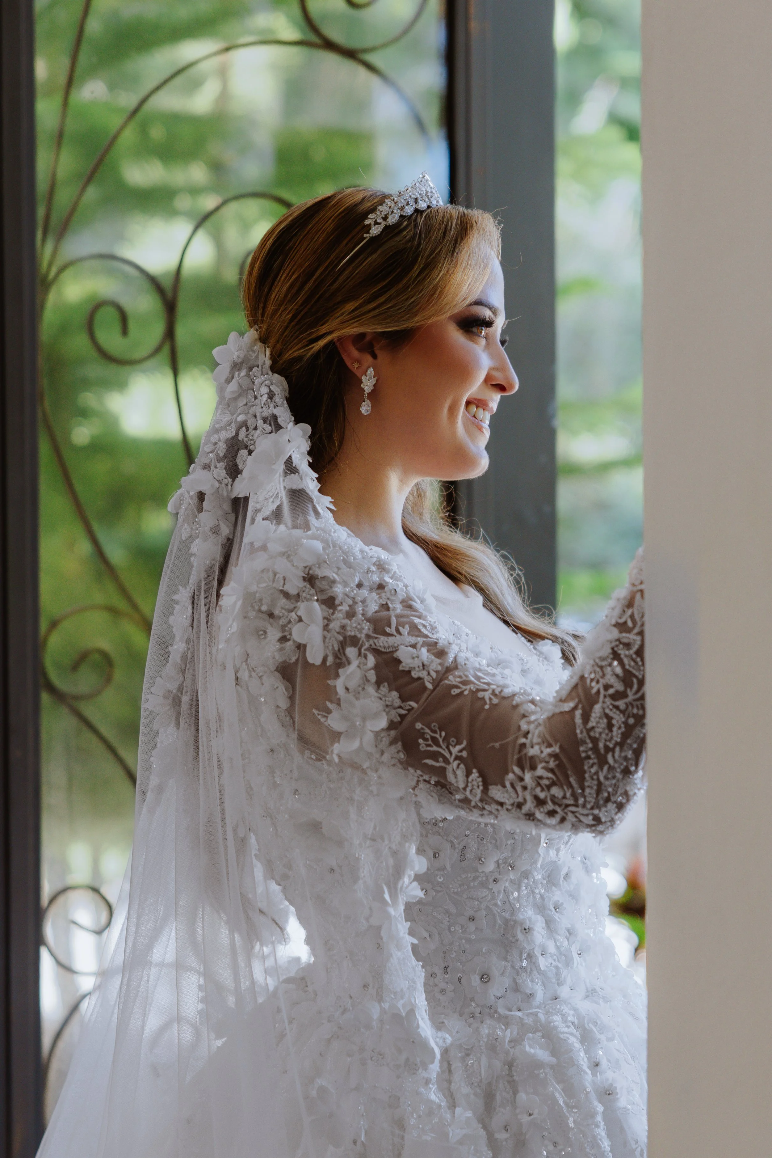 A bride smiling and looking out a window, wearing a lace wedding dress with floral details, a tiara, and earrings.