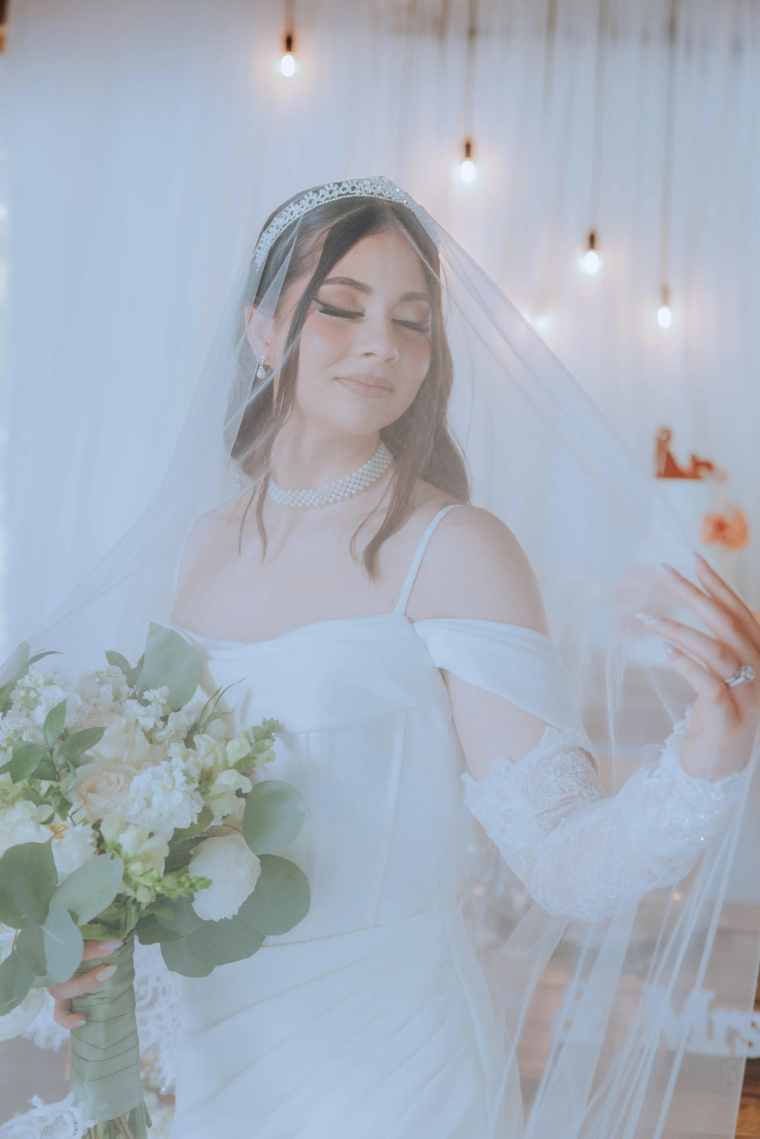 A bride wearing a white wedding dress, veil, pearl necklace, and earrings, holding a bouquet of white roses and greenery, with a soft-focus background featuring hanging lights.