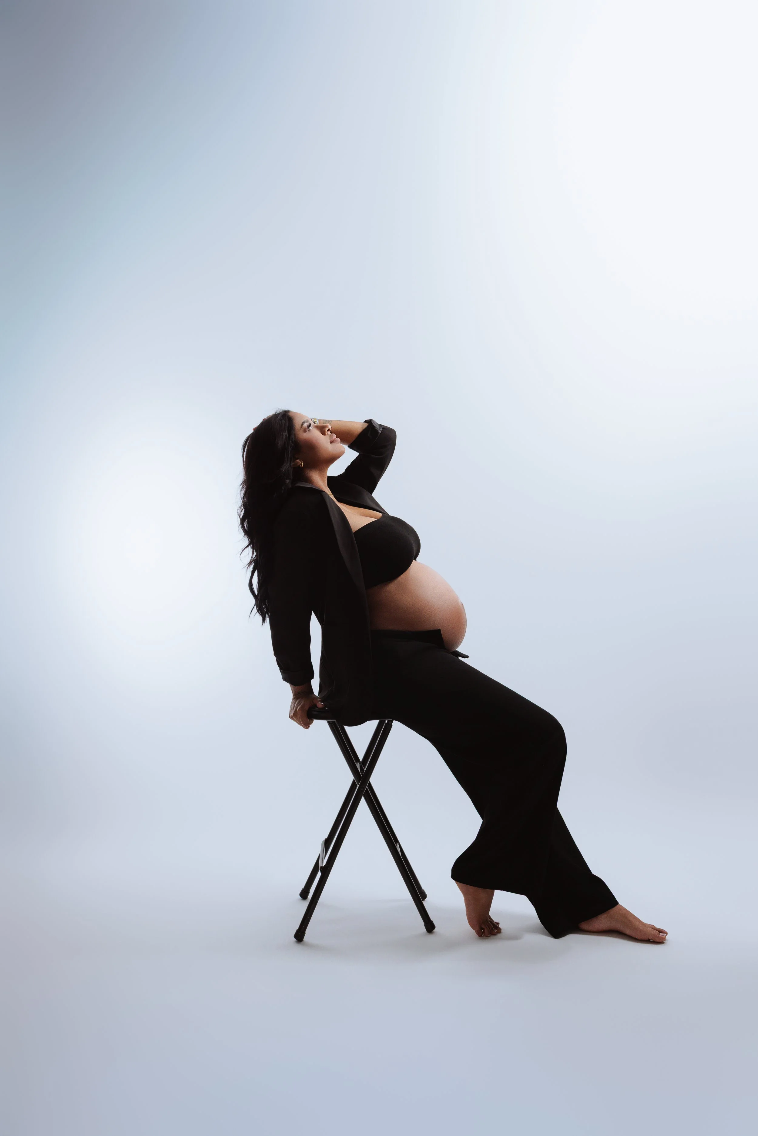 A pregnant woman with long dark hair wearing a black dress and blazer, sitting on a folding chair, posing against a plain light blue background, with one hand behind her head and the other resting on the chair.