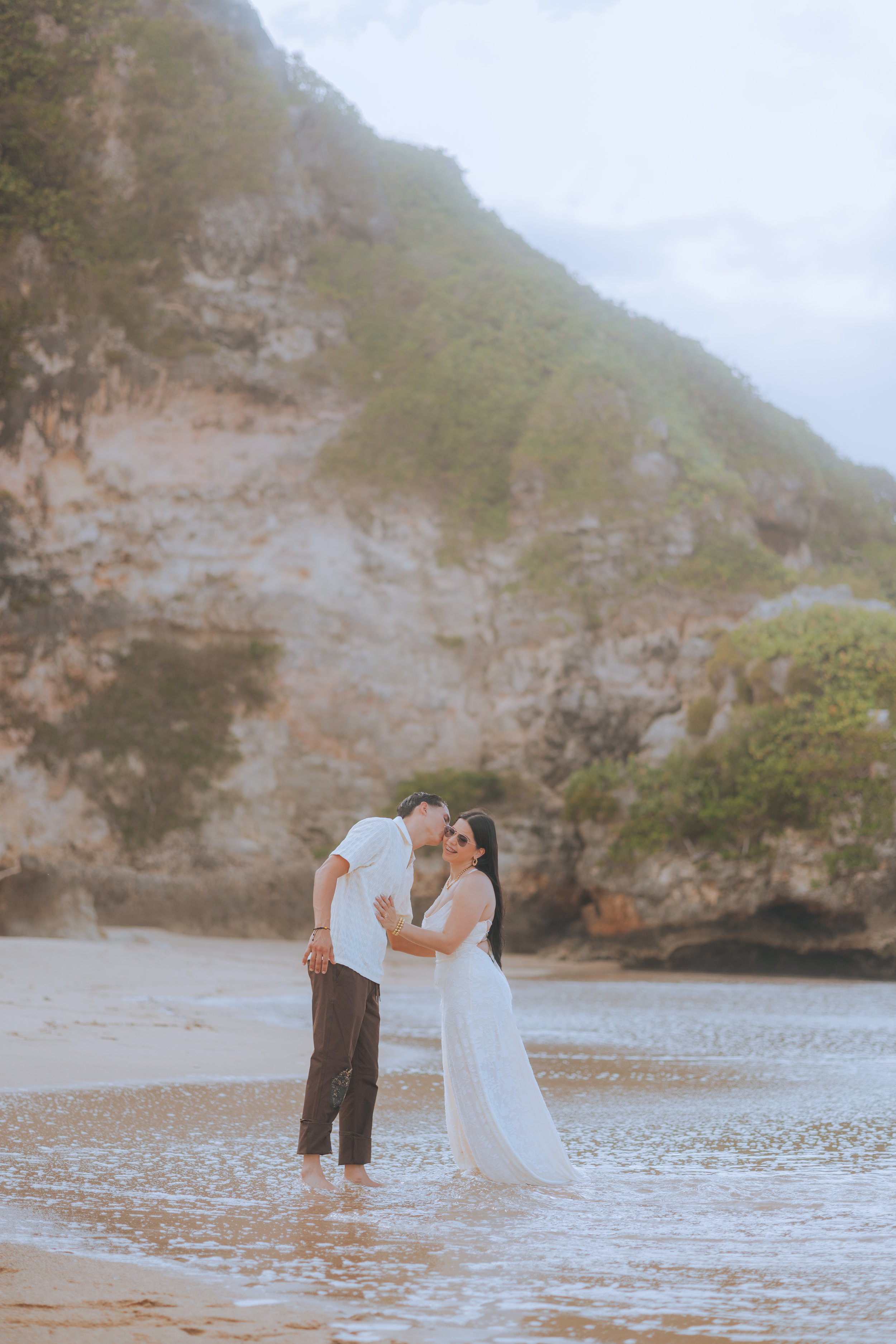 A couple dressed in white standing barefoot in shallow water on a beach, embracing with a rocky green hillside in the background.