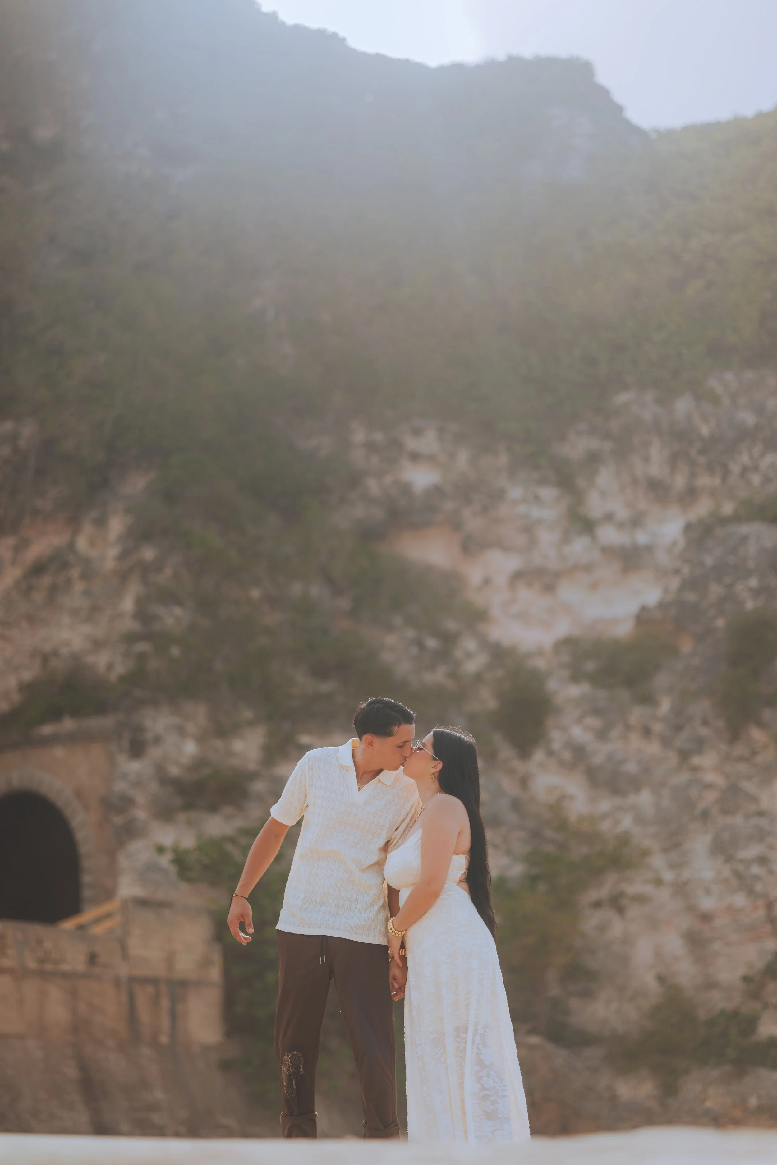 A couple sharing a romantic moment, standing close and leaning in for a kiss, outdoors near an old brick building or ruins.