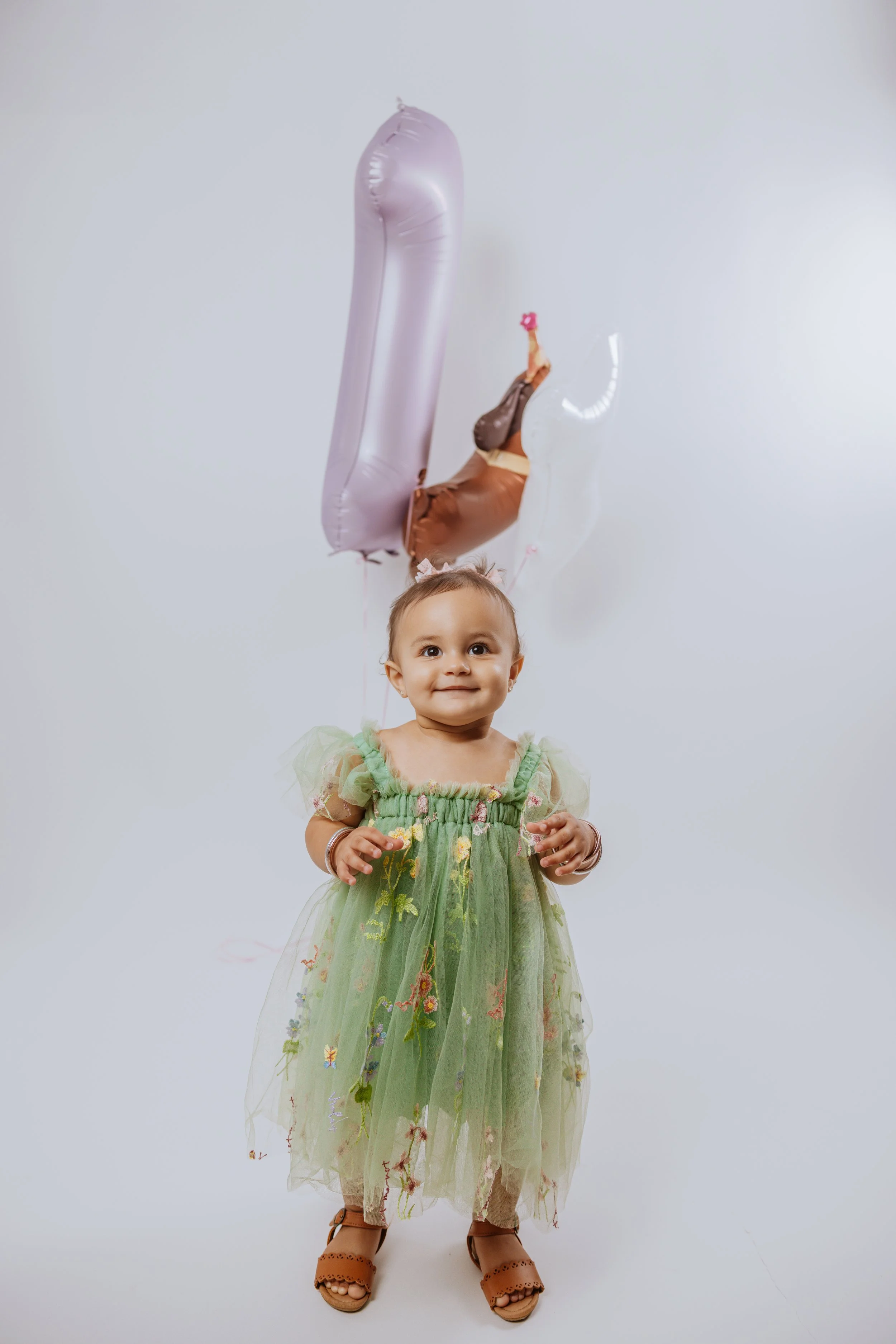 Young girl smiling, wearing a light green dress with floral embroidery, standing in front of a plain white background, holding balloons including a large metallic pink number one and another balloon shaped like a brown animal with pink paws and a flo