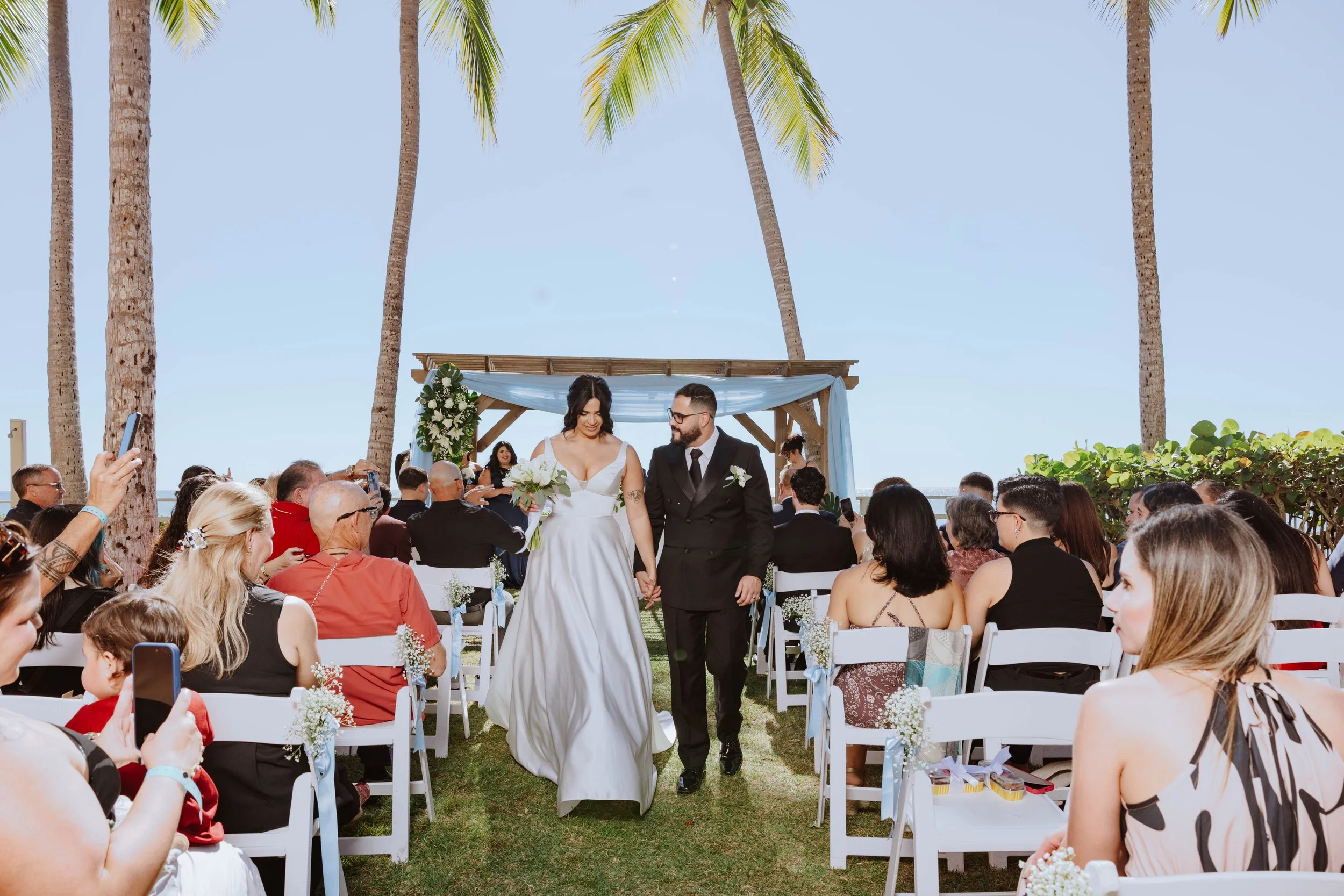 A bride and groom walk hand in hand down the aisle during their outdoor beach wedding ceremony, surrounded by guests seated on white chairs and palm trees in the background.
