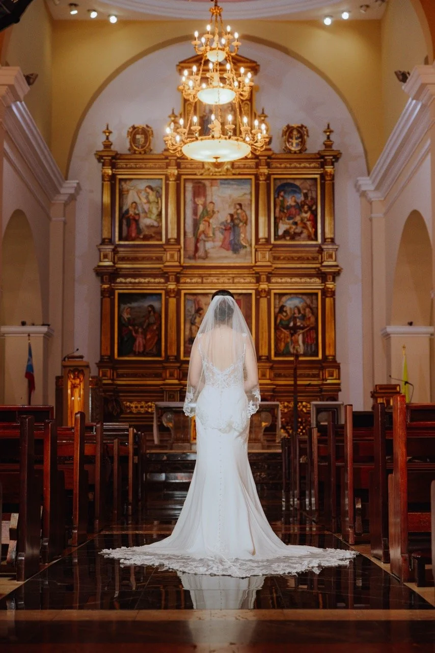A bride in a white wedding dress and veil stands alone in the aisle of a church, facing an ornate altar with religious paintings and chandeliers hanging from the ceiling.