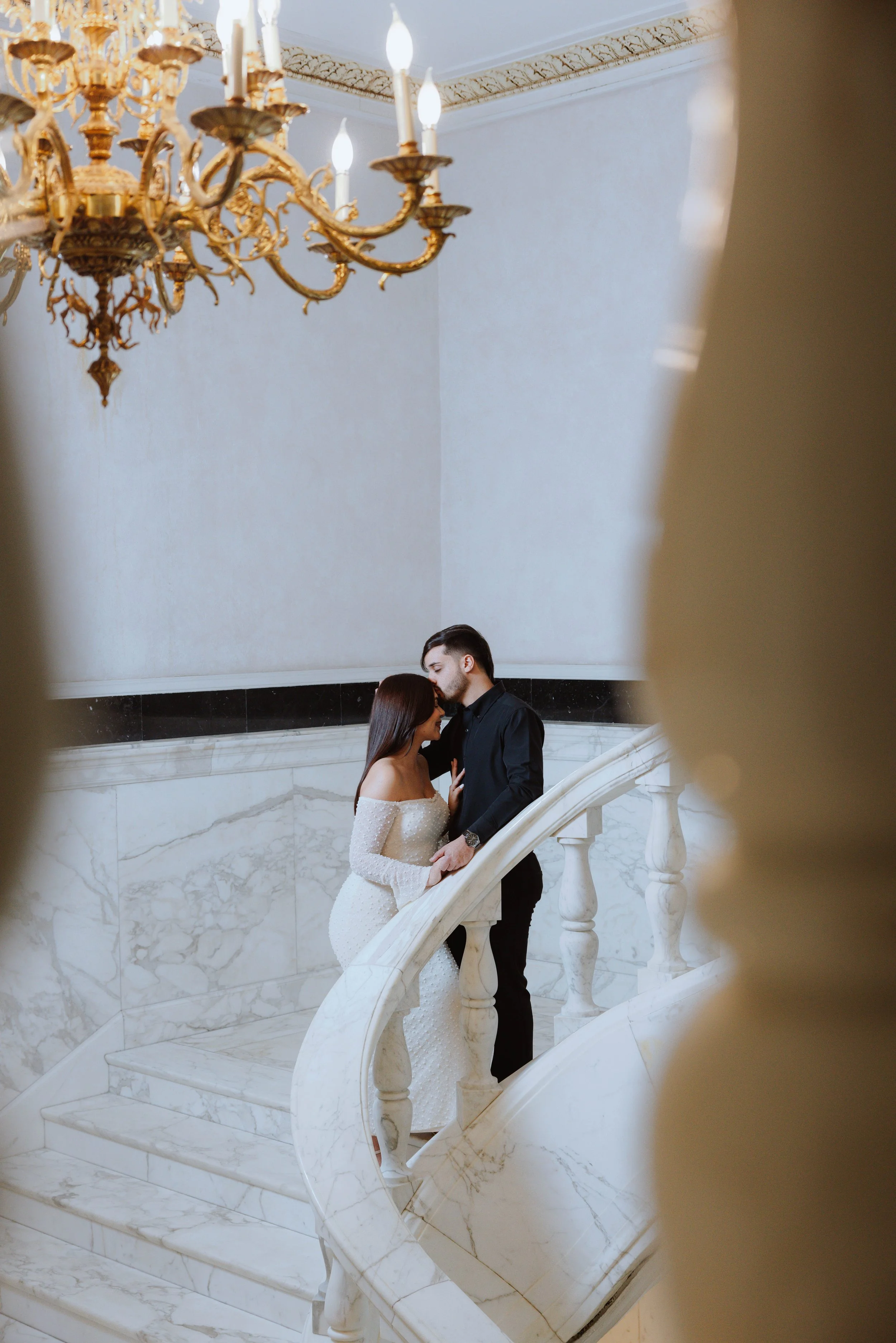 A couple in formal attire, a woman in a white dress and a man in a dark suit, standing on a curved marble staircase inside a grand building with a large gold chandelier hanging from the ceiling.