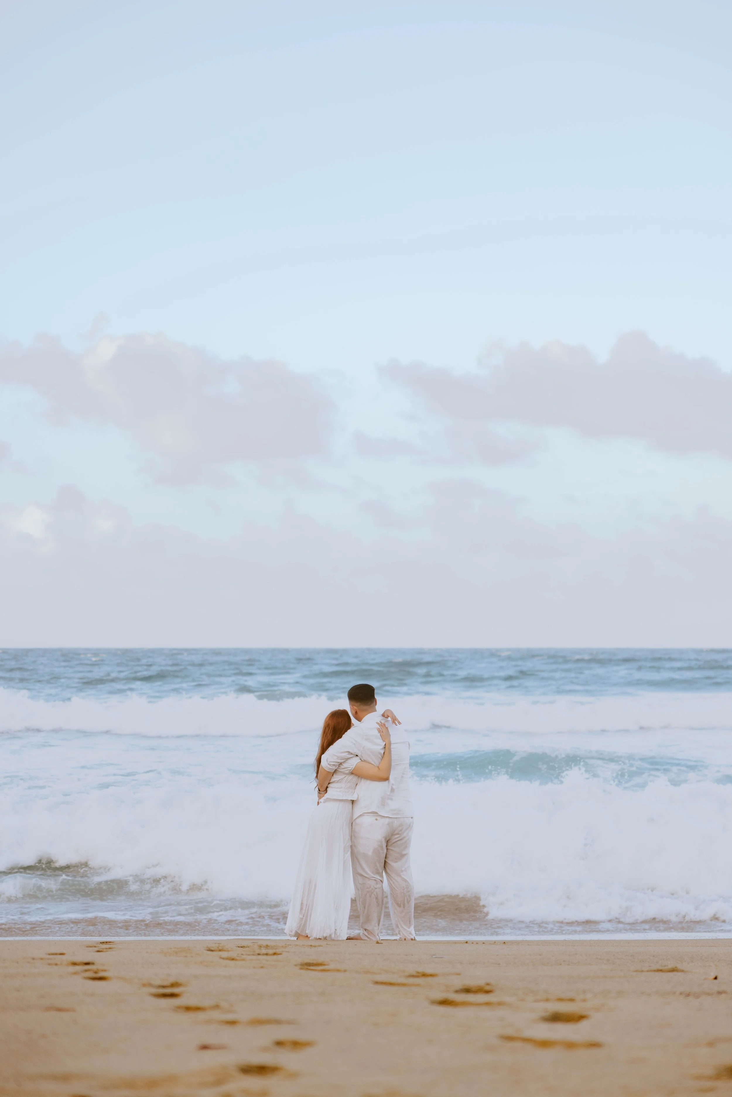 A couple in white clothes hugging on the beach near the ocean during daytime, with cloudy sky above.