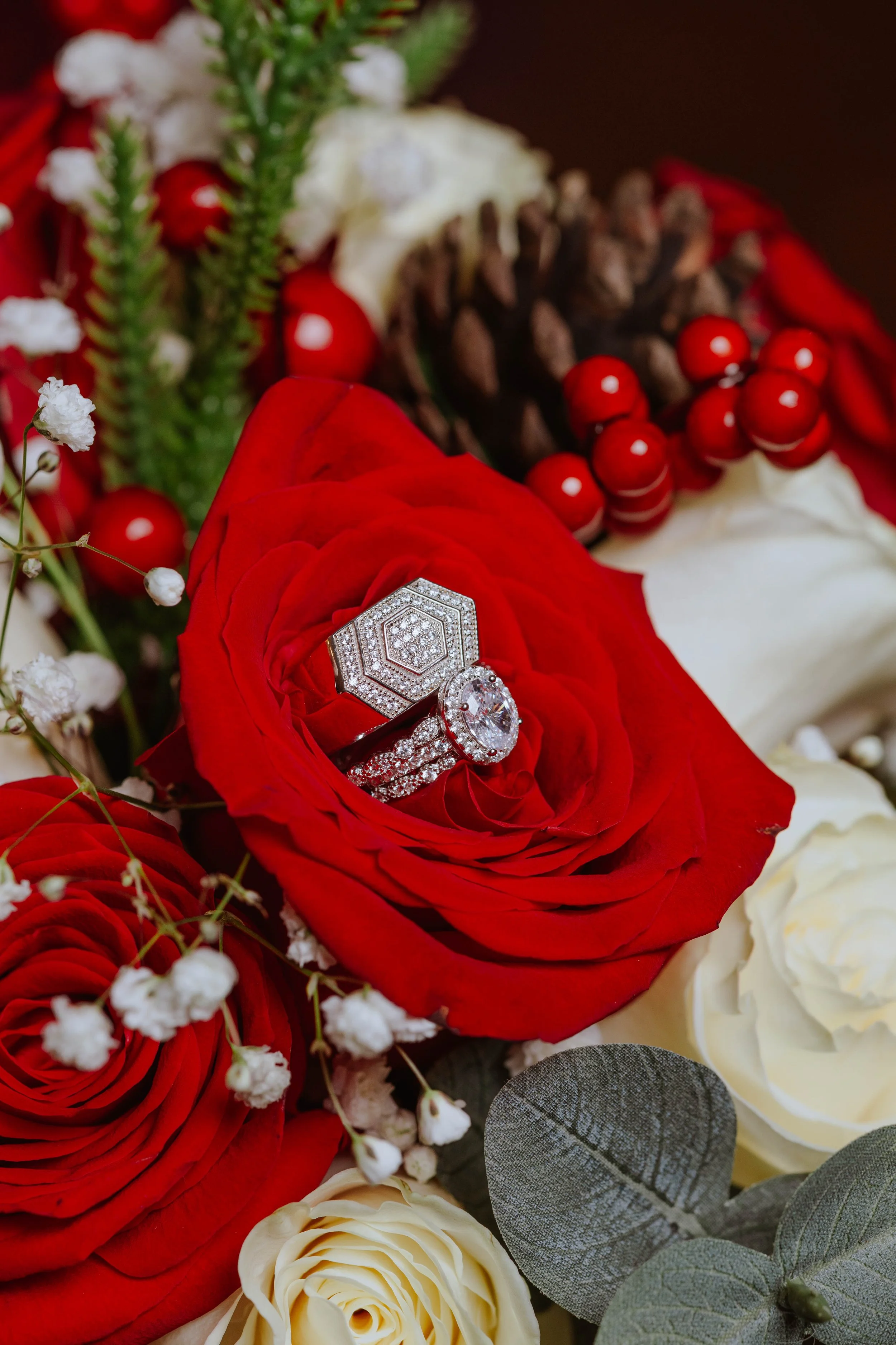 Close-up of a red rose with diamond rings on it, surrounded by white flowers, red berries, pinecones, and eucalyptus leaves.