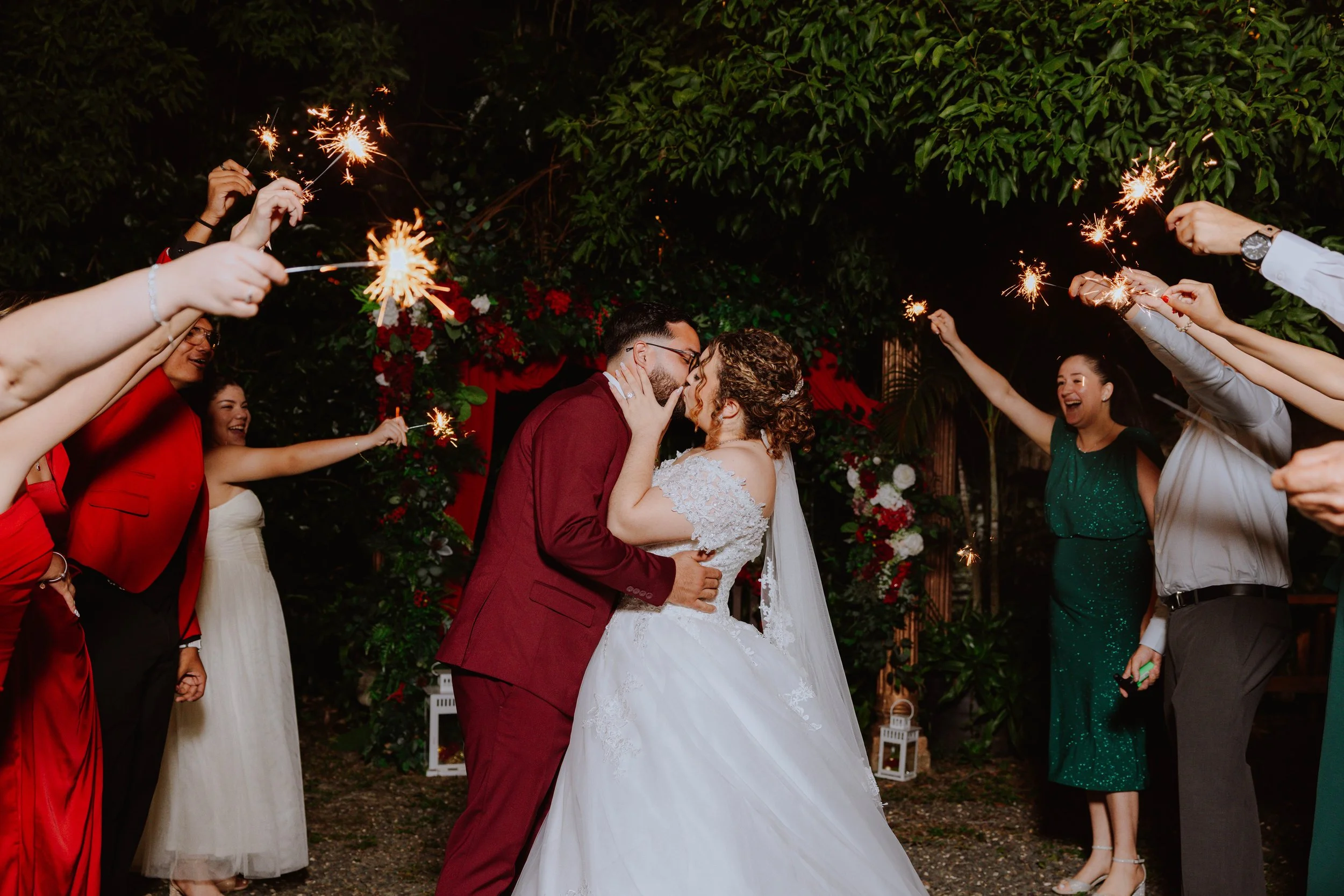 A newlywed couple kissing under sparklers held by friends at night during a wedding celebration outdoors with lush greenery and floral decorations.