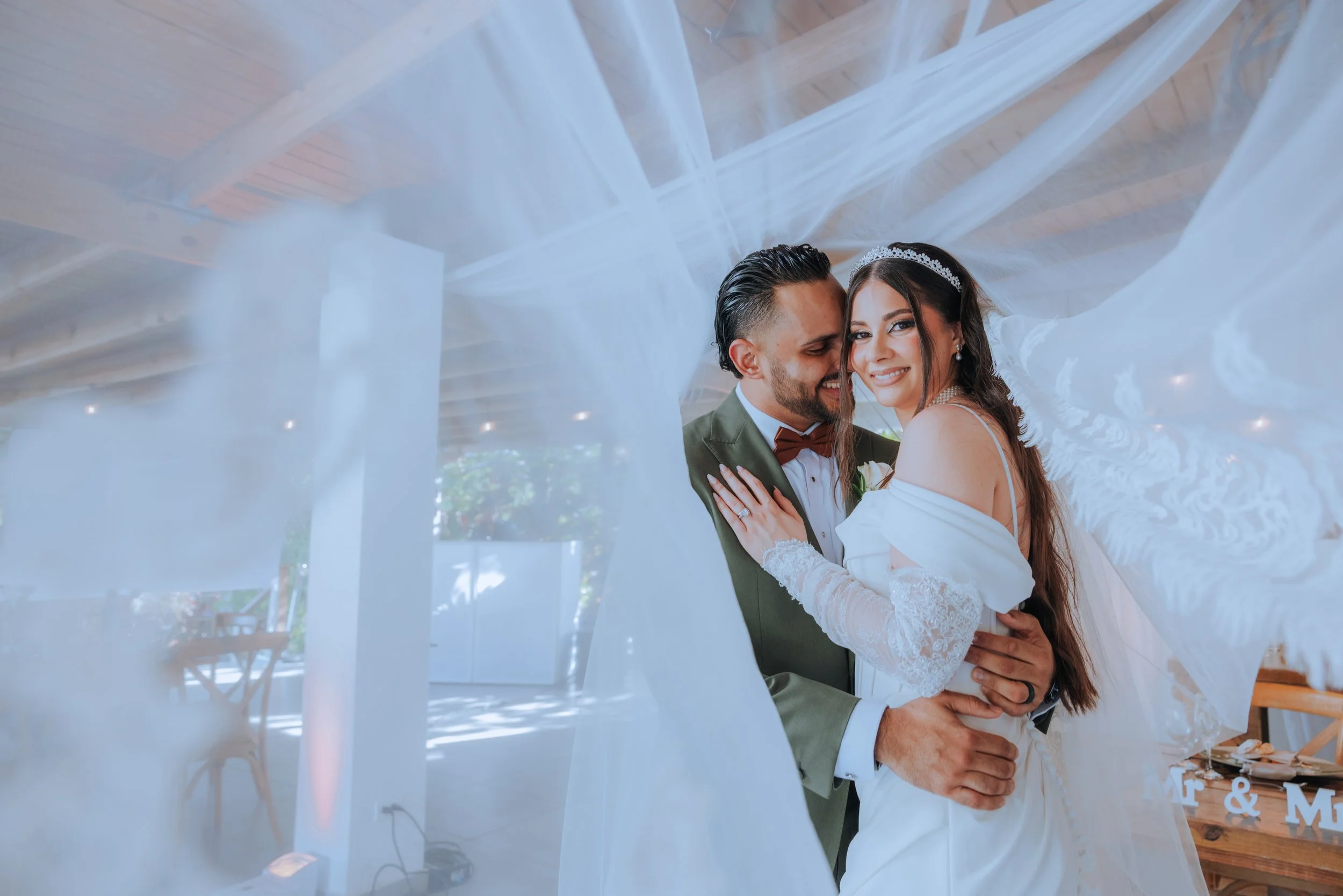 A newly married couple, both smiling and embracing, standing under a white canopy at their wedding reception. The bride is wearing a tiara and pearl earrings, and the groom is in a green suit with a brown bow tie.