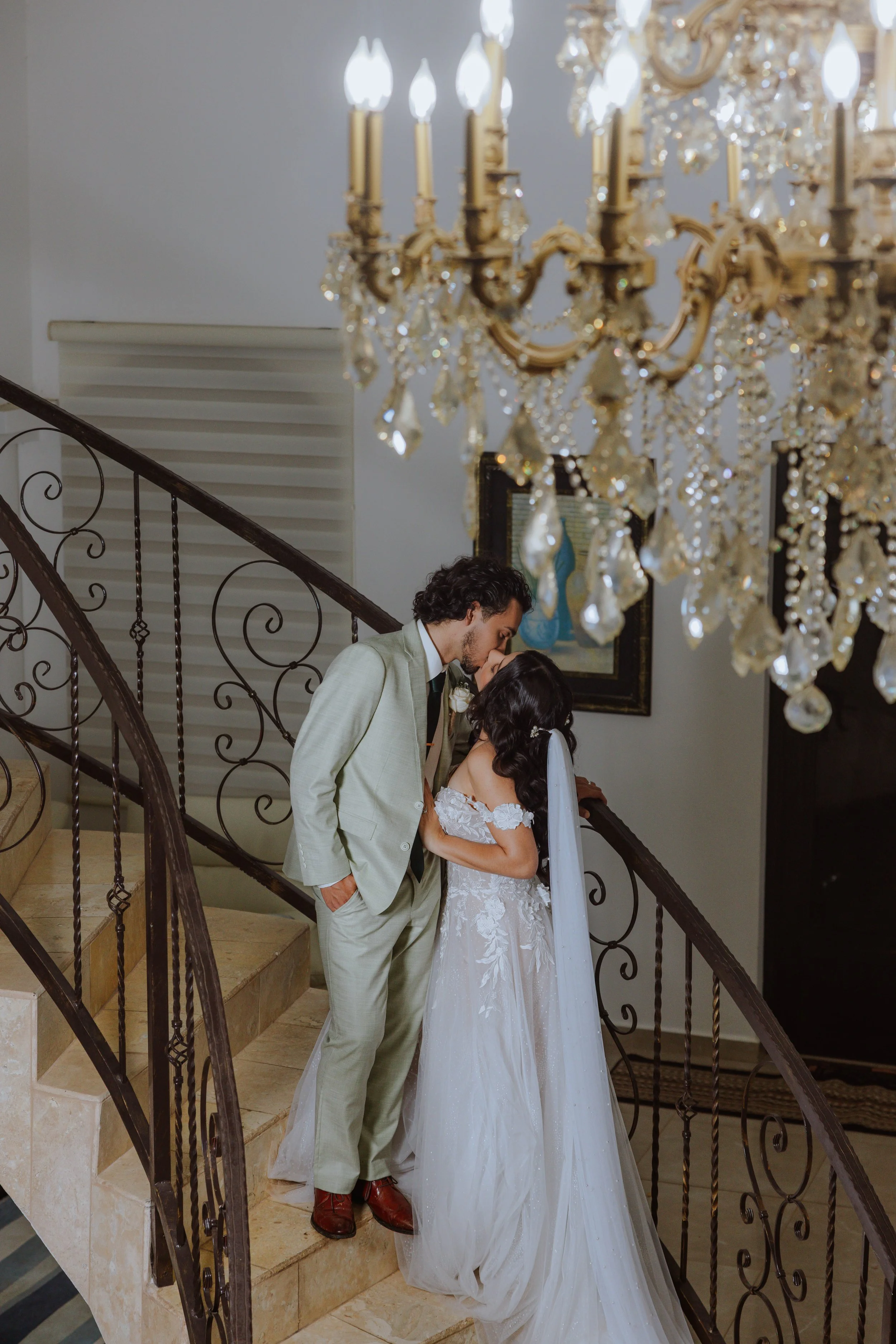 A couple in wedding attire standing on a staircase, about to kiss, with a chandelier hanging above them.