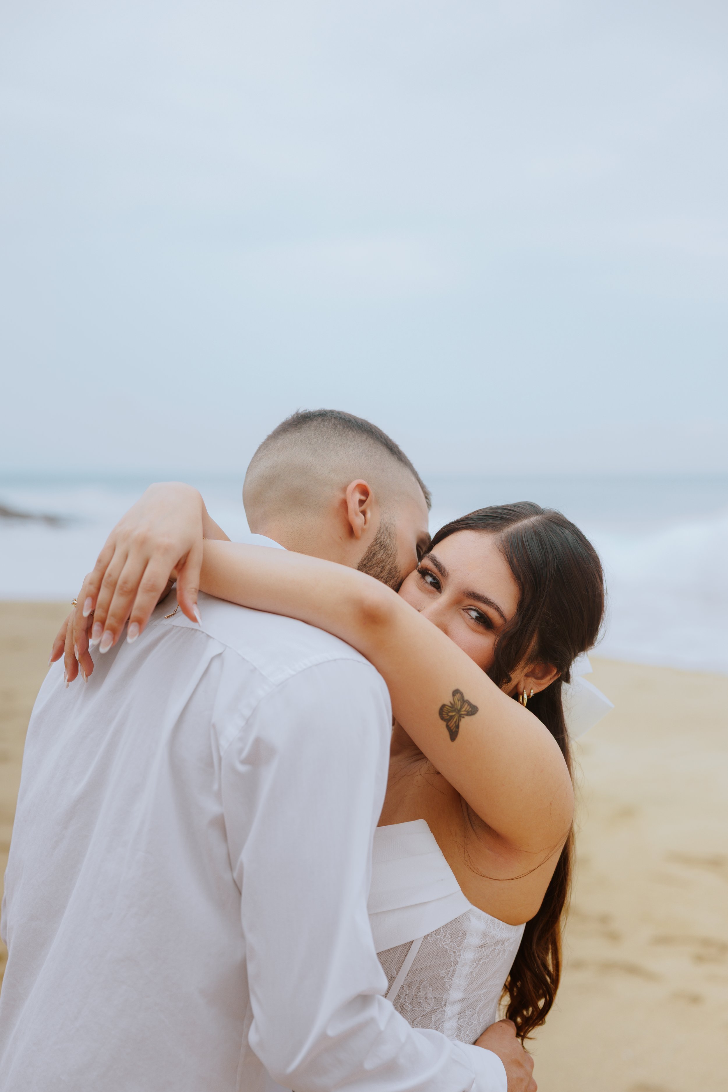 A couple sharing an intimate hug on a beach, with the man kissing the woman on the forehead and both smiling, during daytime.