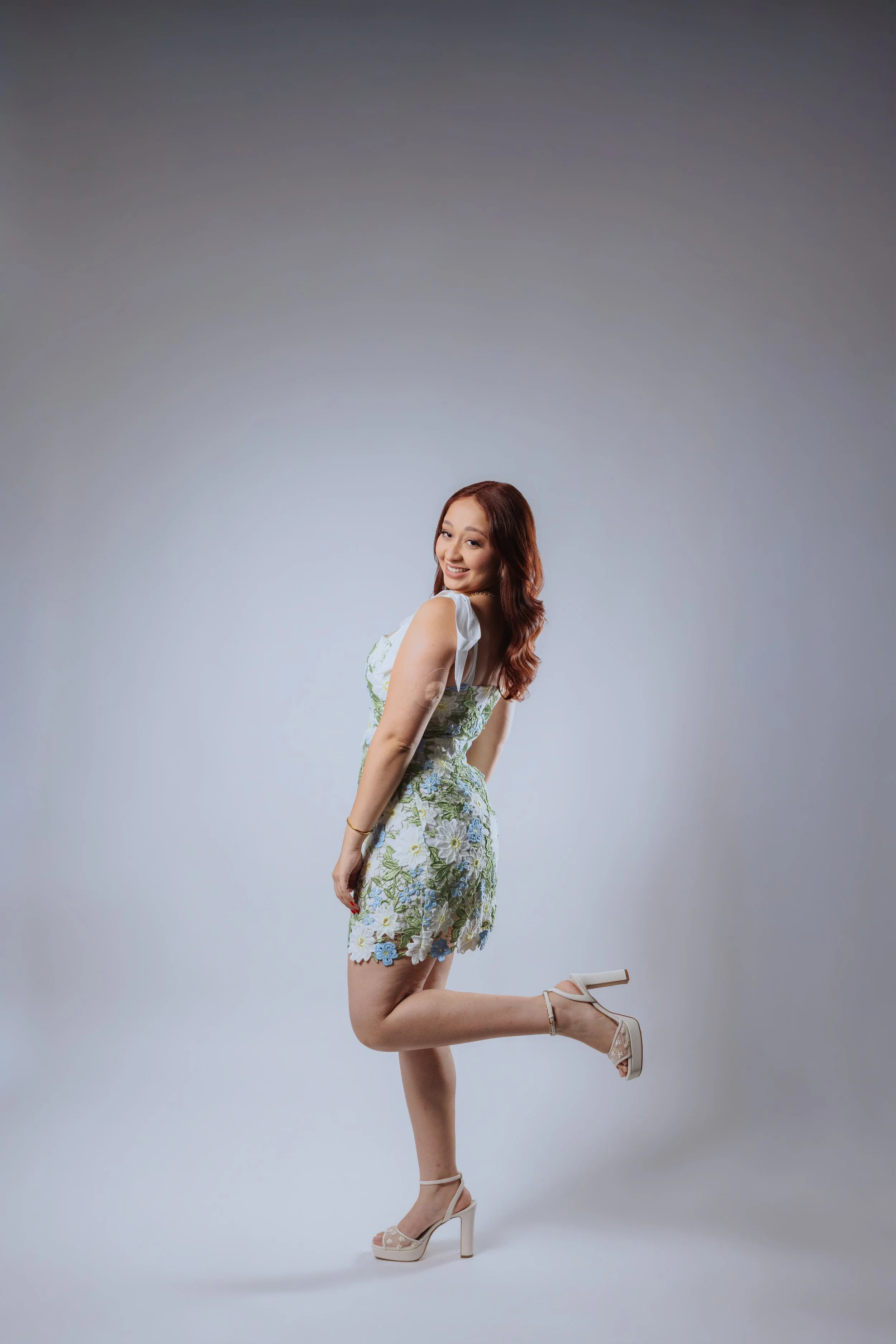 A woman with long brown hair wearing a floral dress with sheer shoes, smiling and kneeling on one knee against a plain gray background.