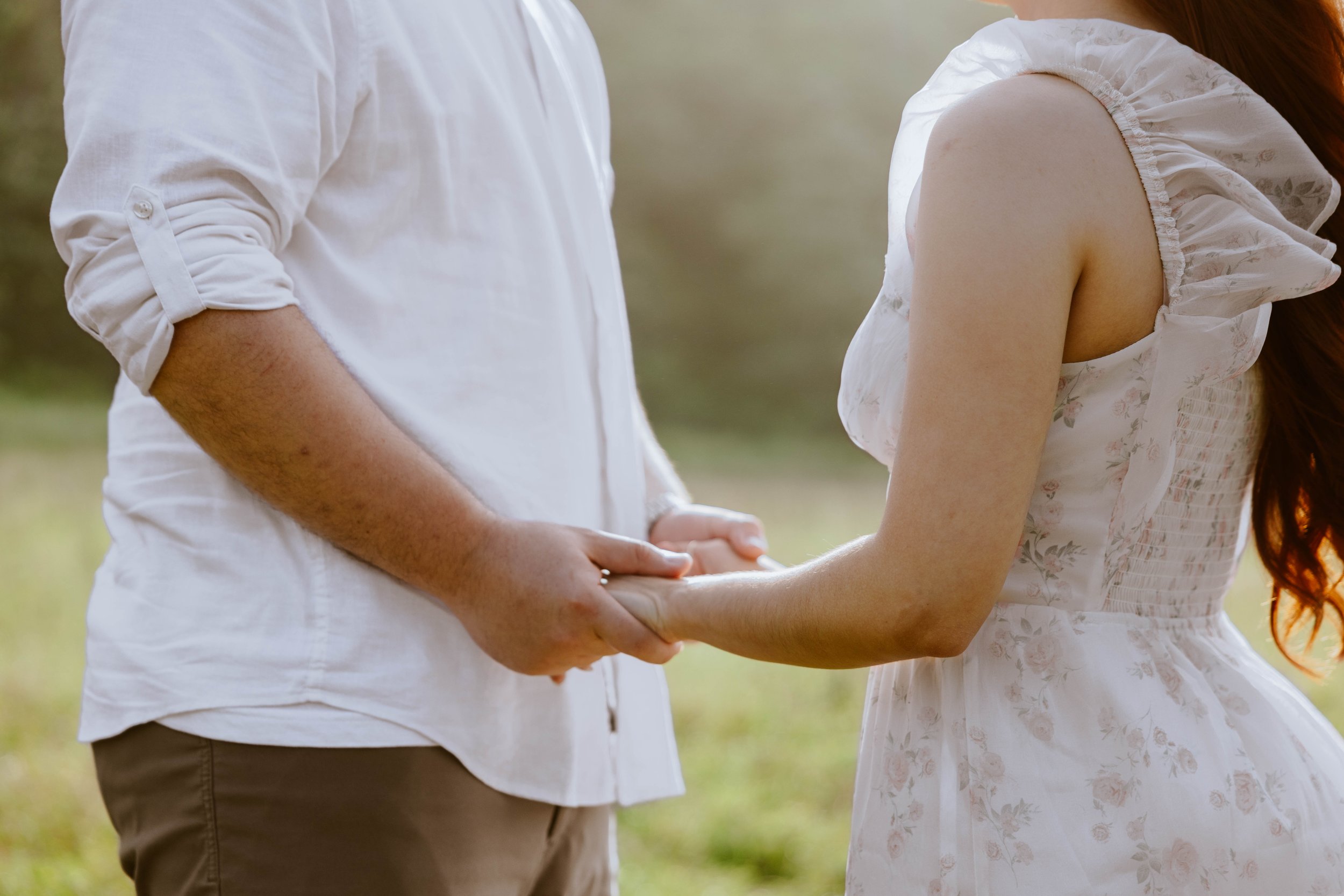 A man and a woman holding hands outdoors, with the woman wearing a white dress with ruffled sleeves.