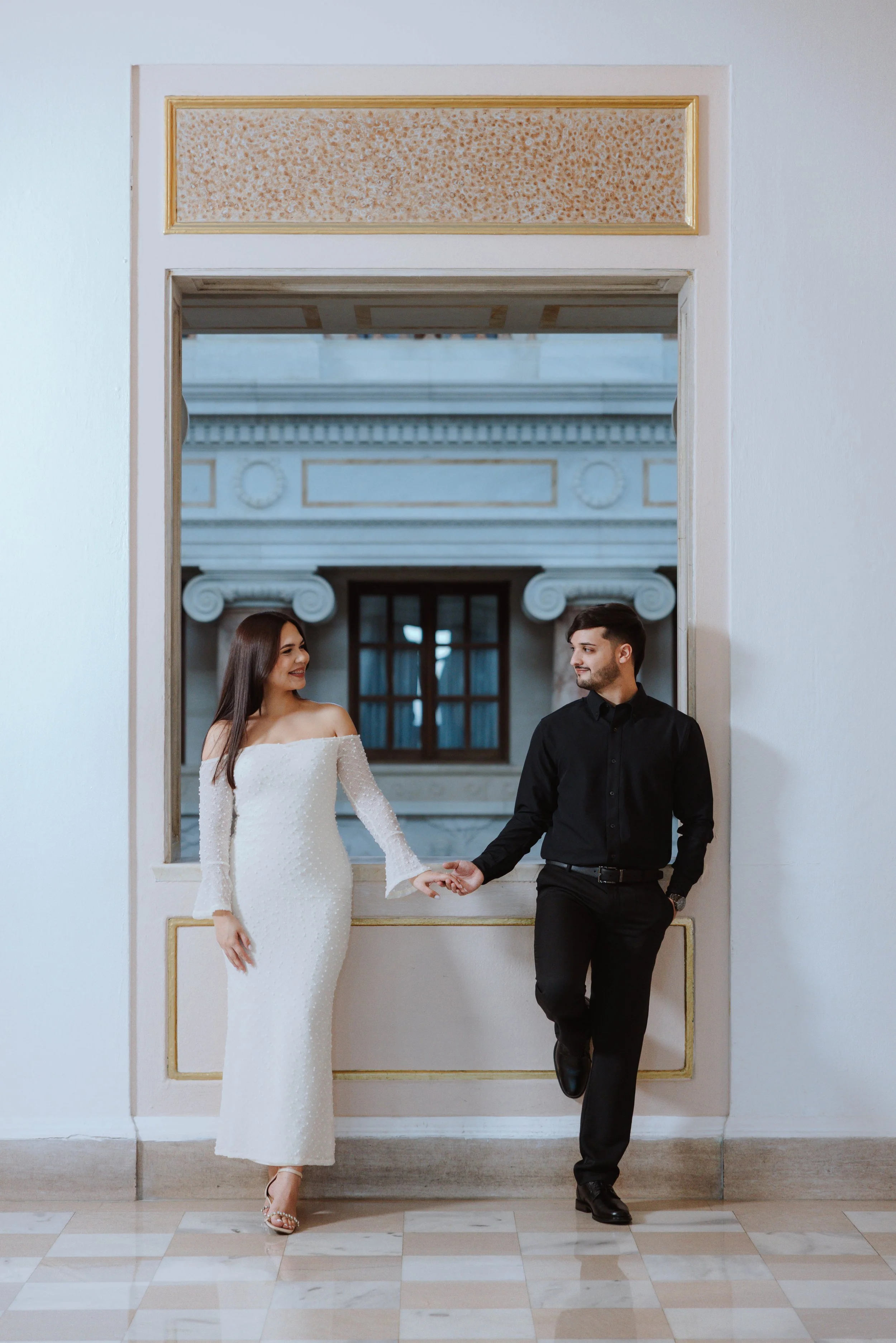 A woman in a white wedding dress and a man in a black suit holding hands and smiling at each other inside a building with ornate architecture.