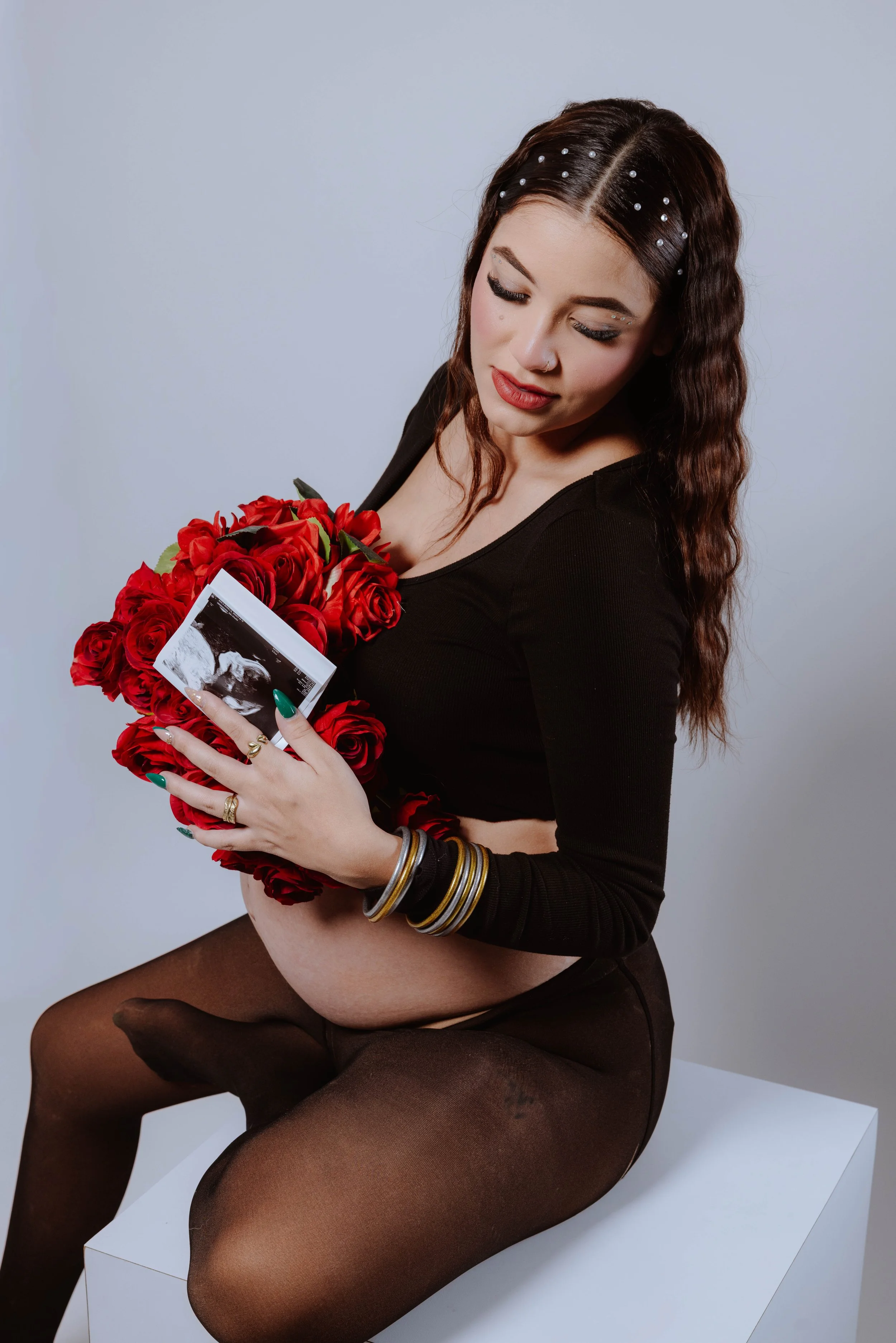 A woman with long wavy brown hair and pearl hair accessories is sitting on a white block, wearing a black top and sheer black tights. She is holding a bouquet of red roses and an ultrasound photo, and has multiple bracelets on her wrists.