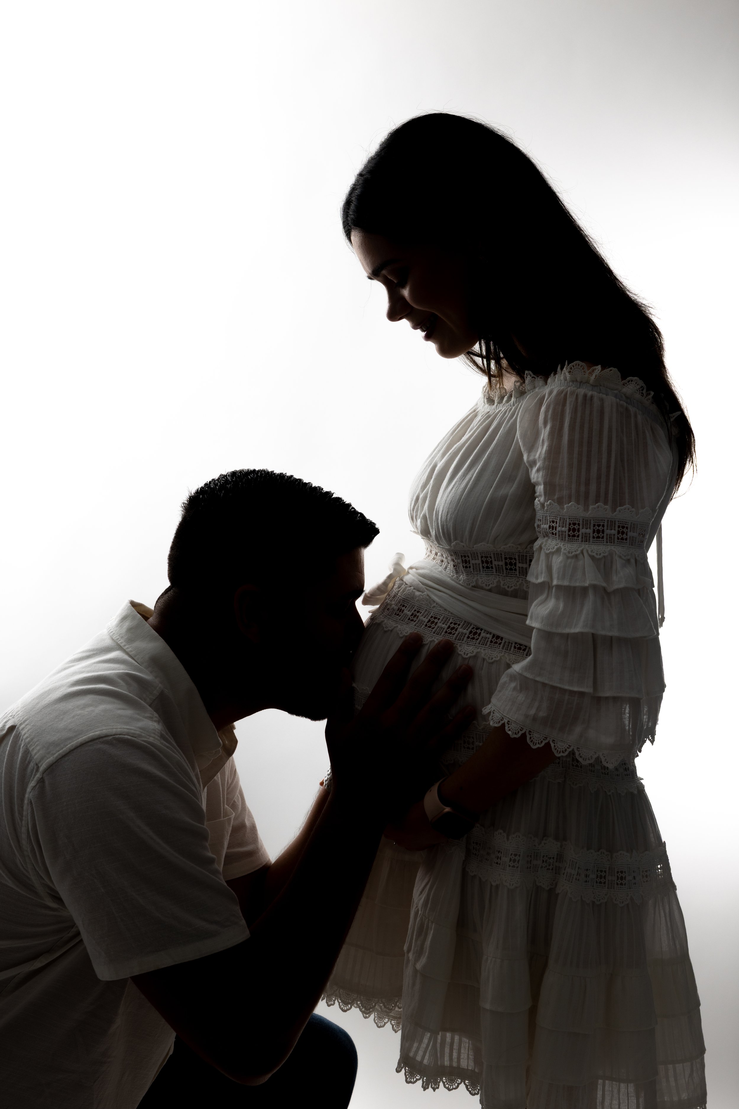 Silhouette of a man kneeling with hands on a pregnant woman's belly, who is smiling and looking down, both in profile against a bright background.