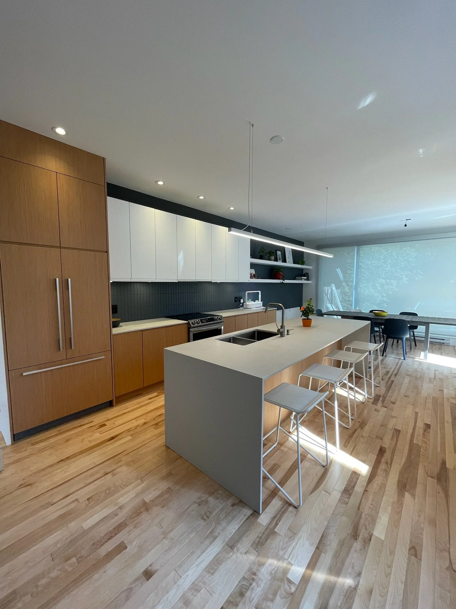 Modern kitchen with wooden cabinets, white and dark-colored shelves, a kitchen island with a sink, barstools, a dining table with chairs, and large windows with natural light.