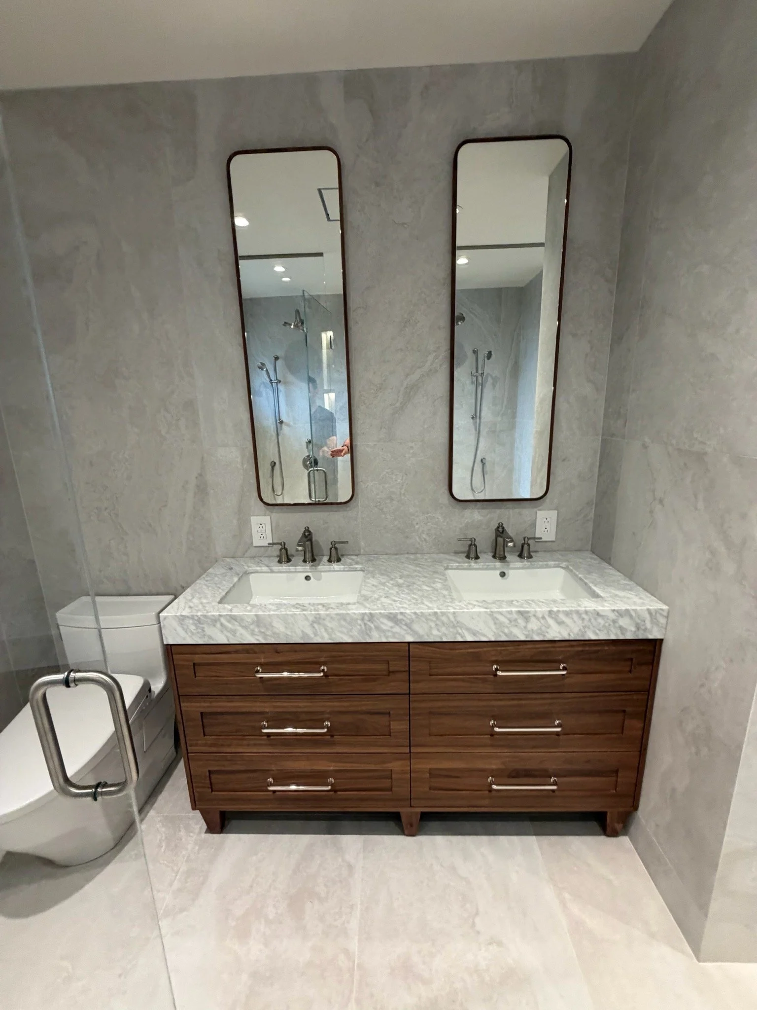 Bathroom vanity with a marble countertop, double sinks, and two framed mirrors, with a toilet to the left and tiled walls and floor.