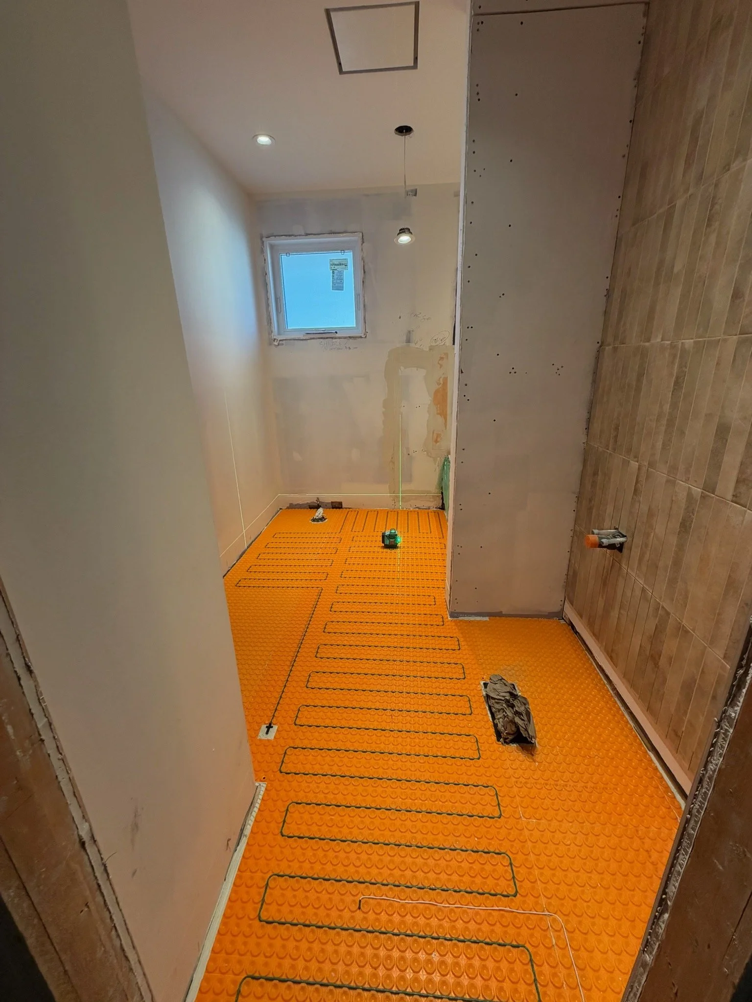 Room under renovation with exposed drywall, a small window, hardwood and tile floors, and an orange mat with black heating cables laid out for installation.