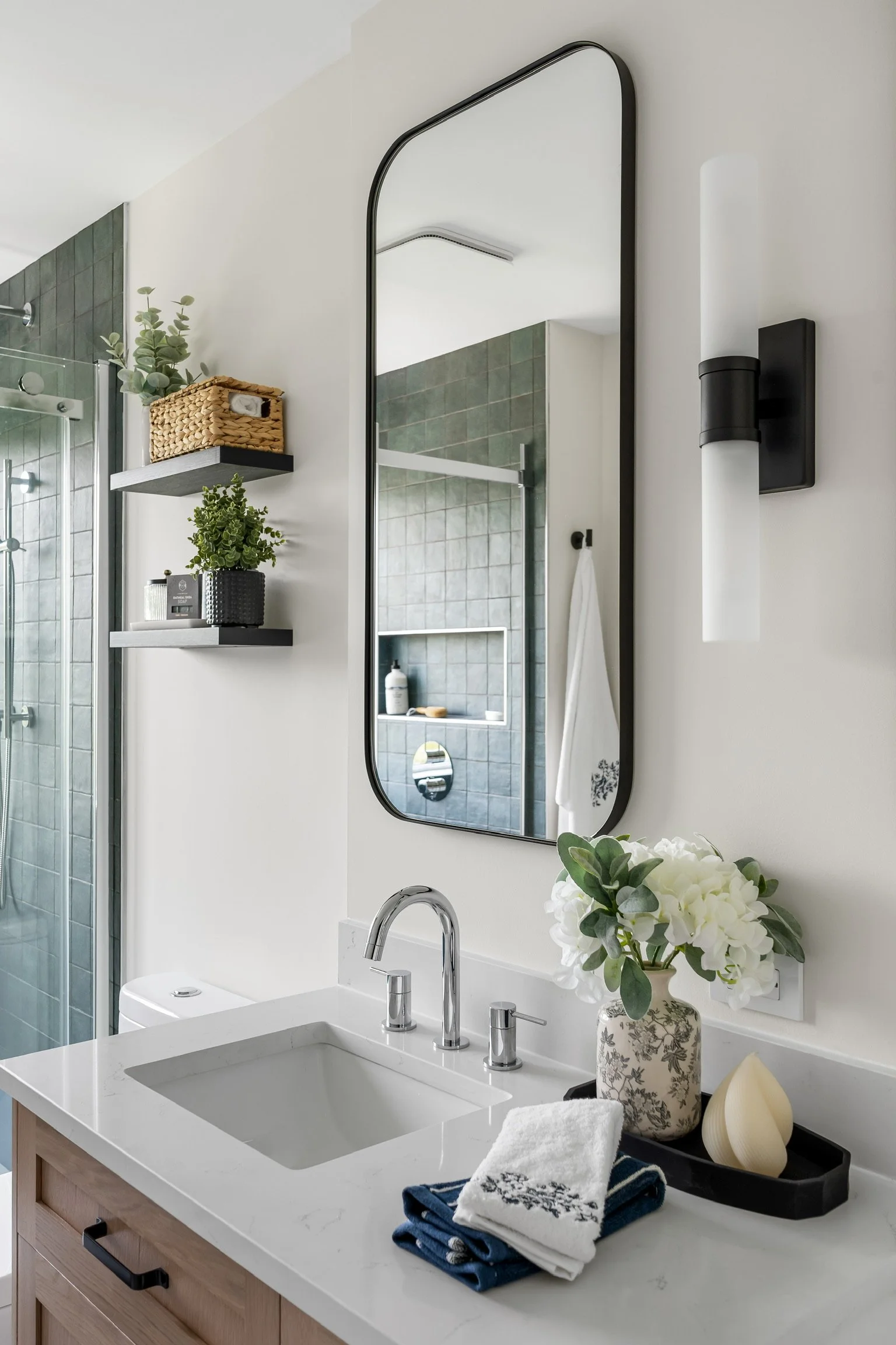 Bathroom vanity with a white countertop, silver faucet, and a vase of white flowers. Two folded towels, one white with gray embroidery and one navy, are placed on a black tray next to the sink. A large mirror and a black wall sconce are above the van