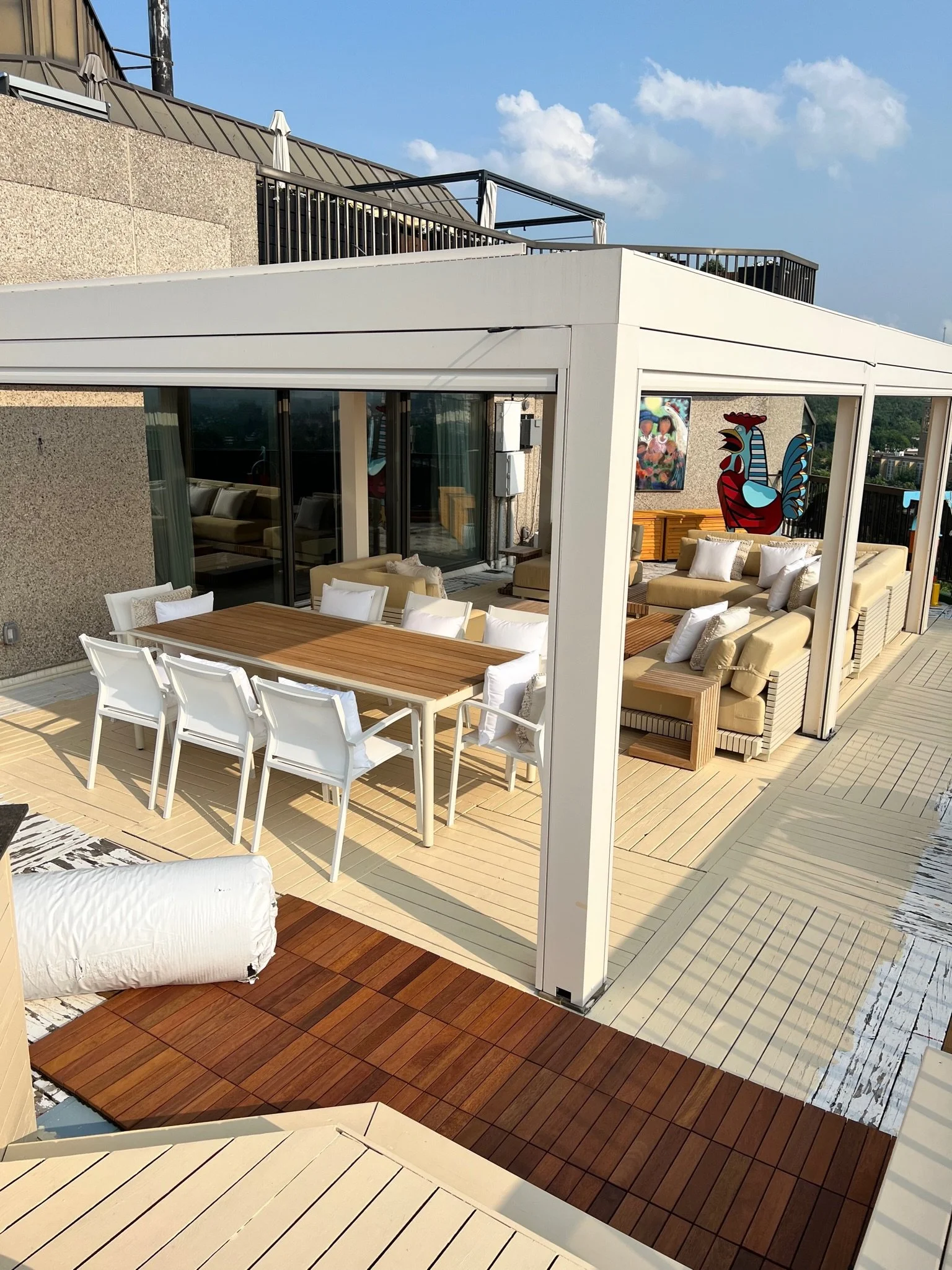A rooftop terrace with wooden and white flooring, outdoor seating, a white canopy structure, a colorful rooster mural, and a city view in the background.