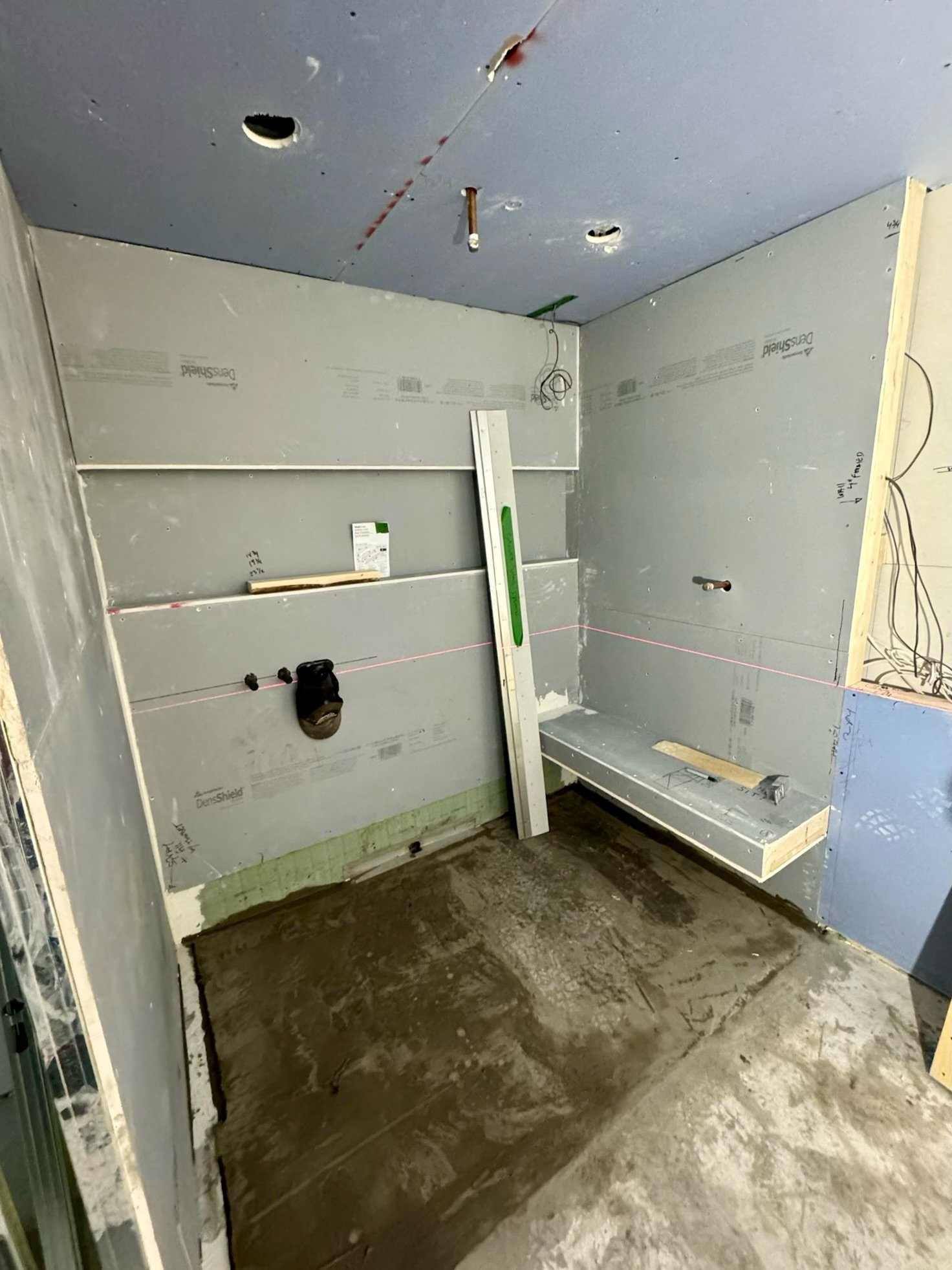 Construction of a bathroom with gray drywall ceiling and walls, some wires hanging, a level tool standing upright, and a wooden frame on the floor, with a concrete slab beneath.