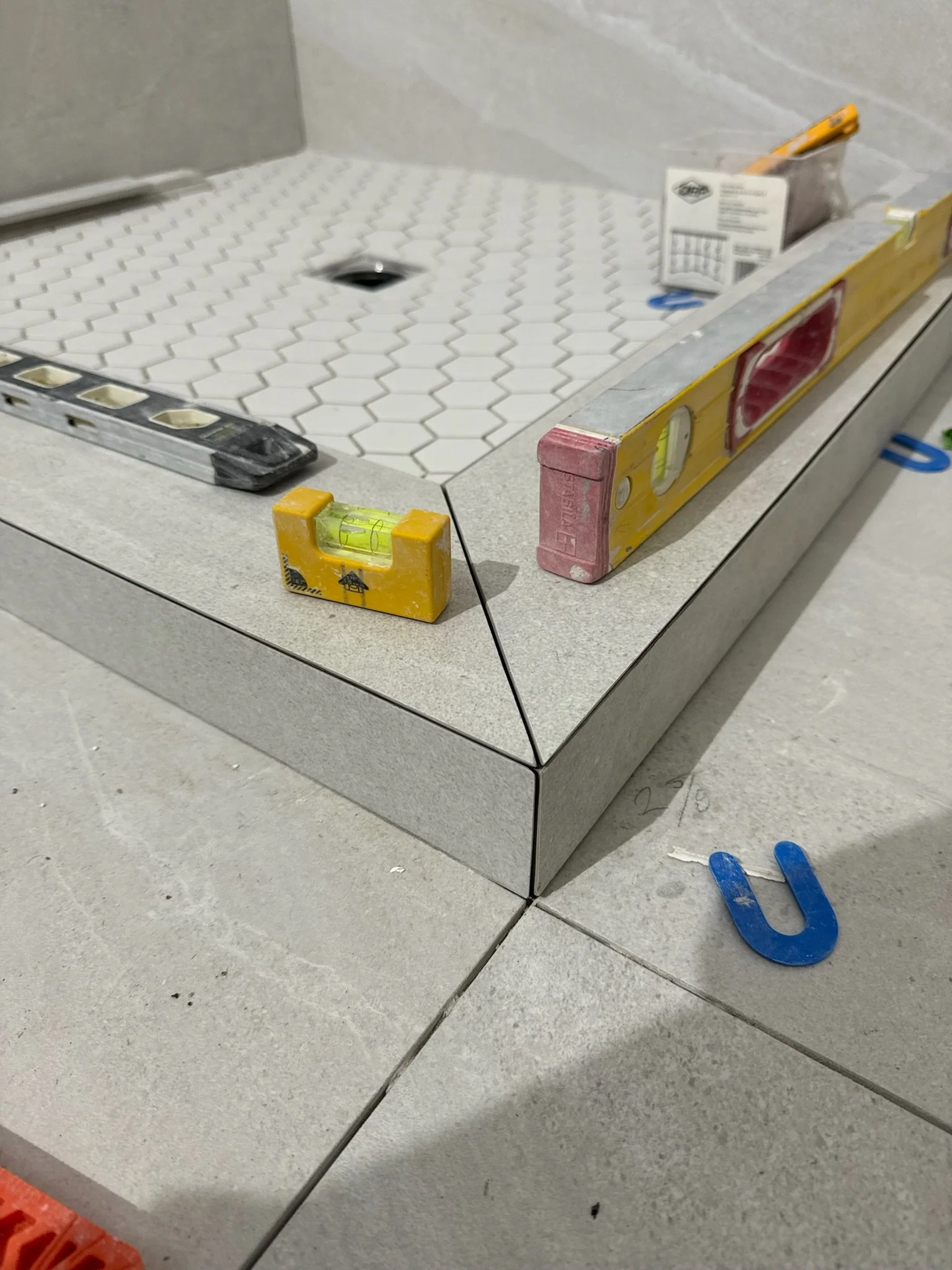 Close-up of a construction site showing tile flooring installation with construction tools including a level, blue spacers, and a pencil on the surface.