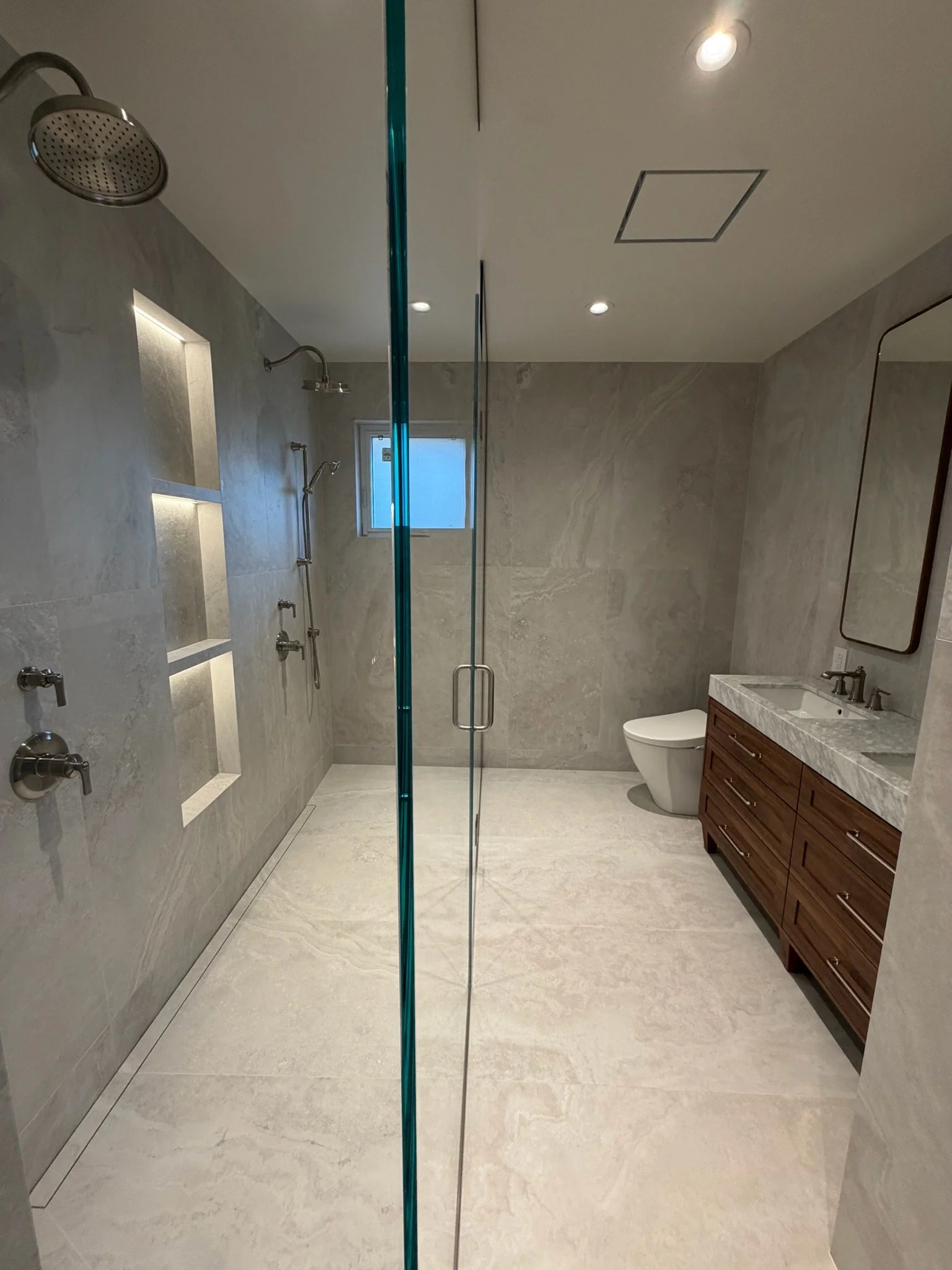 Modern bathroom with large shower area featuring multiple shower heads, a small window, and a vanity with a marble countertop and a mirror.