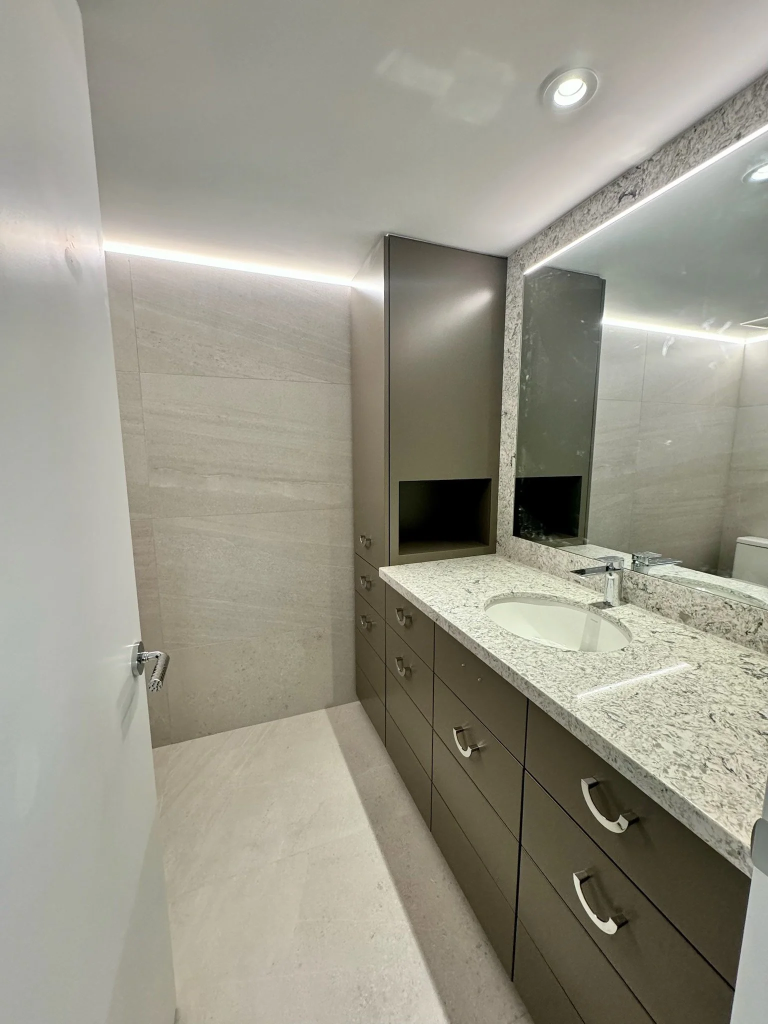 Modern bathroom with beige tiled walls and floor, a long granite countertop with an oval sink, gray cabinetry with drawers and silver handles, a large mirror, and recessed ceiling lights.