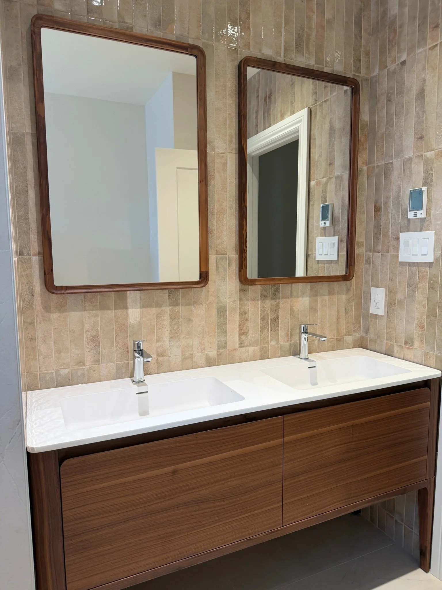 Double vanity bathroom with two rectangular mirrors with wooden frames, beige tiled wall, twin sinks, and a wooden cabinet below the sinks.
