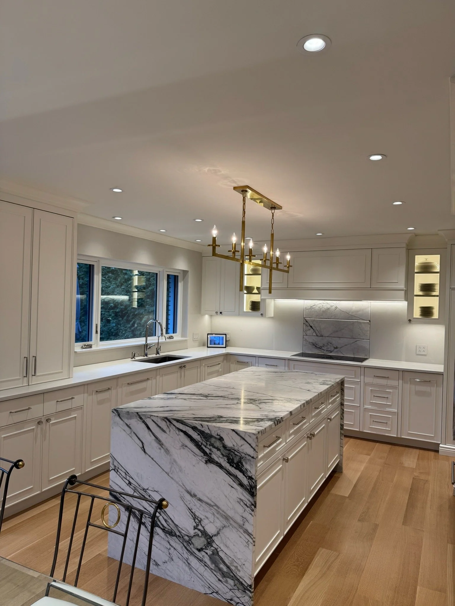 A modern kitchen with white cabinetry, a marble island with black and gray veining, large window, and a gold chandelier with six candle-style lights.