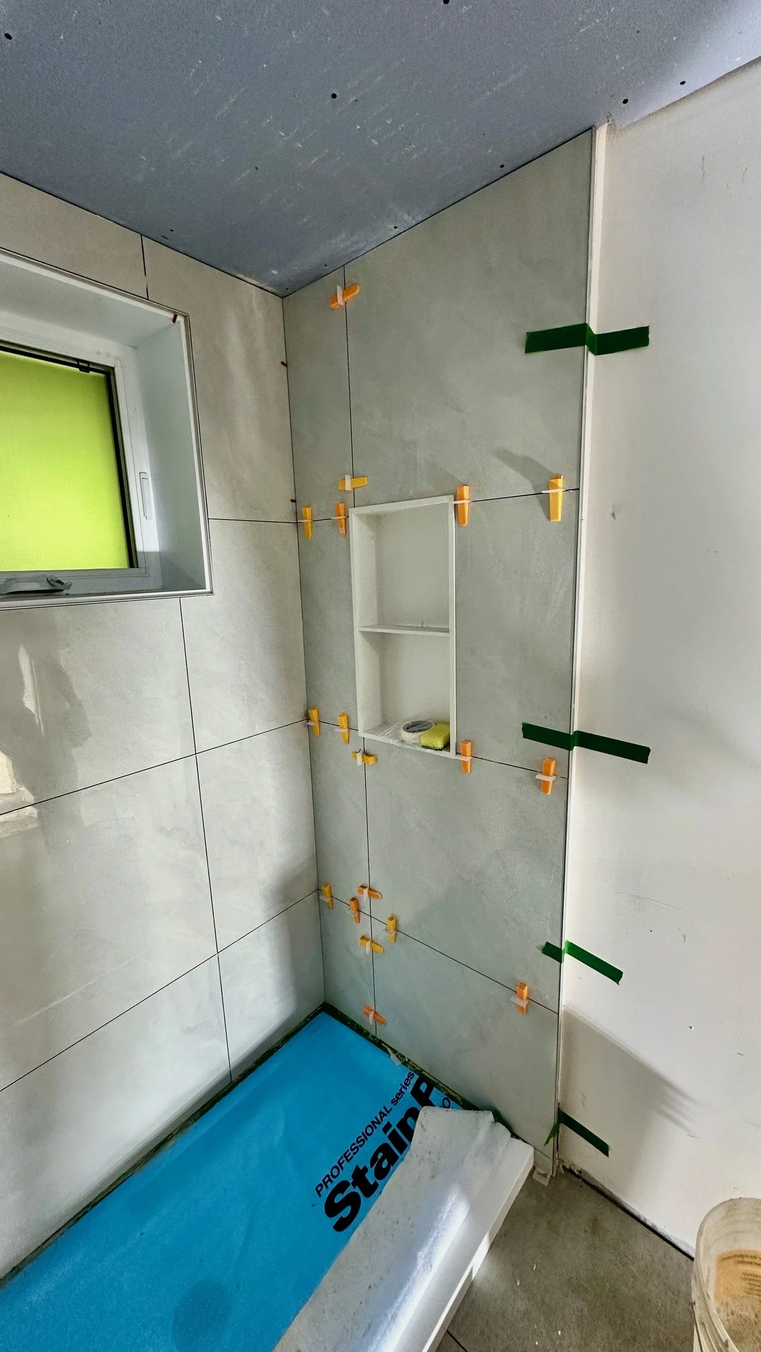 A bathroom under renovation with tiled walls, a window, and a partially installed wall shelf secured with orange and green tile spacers.