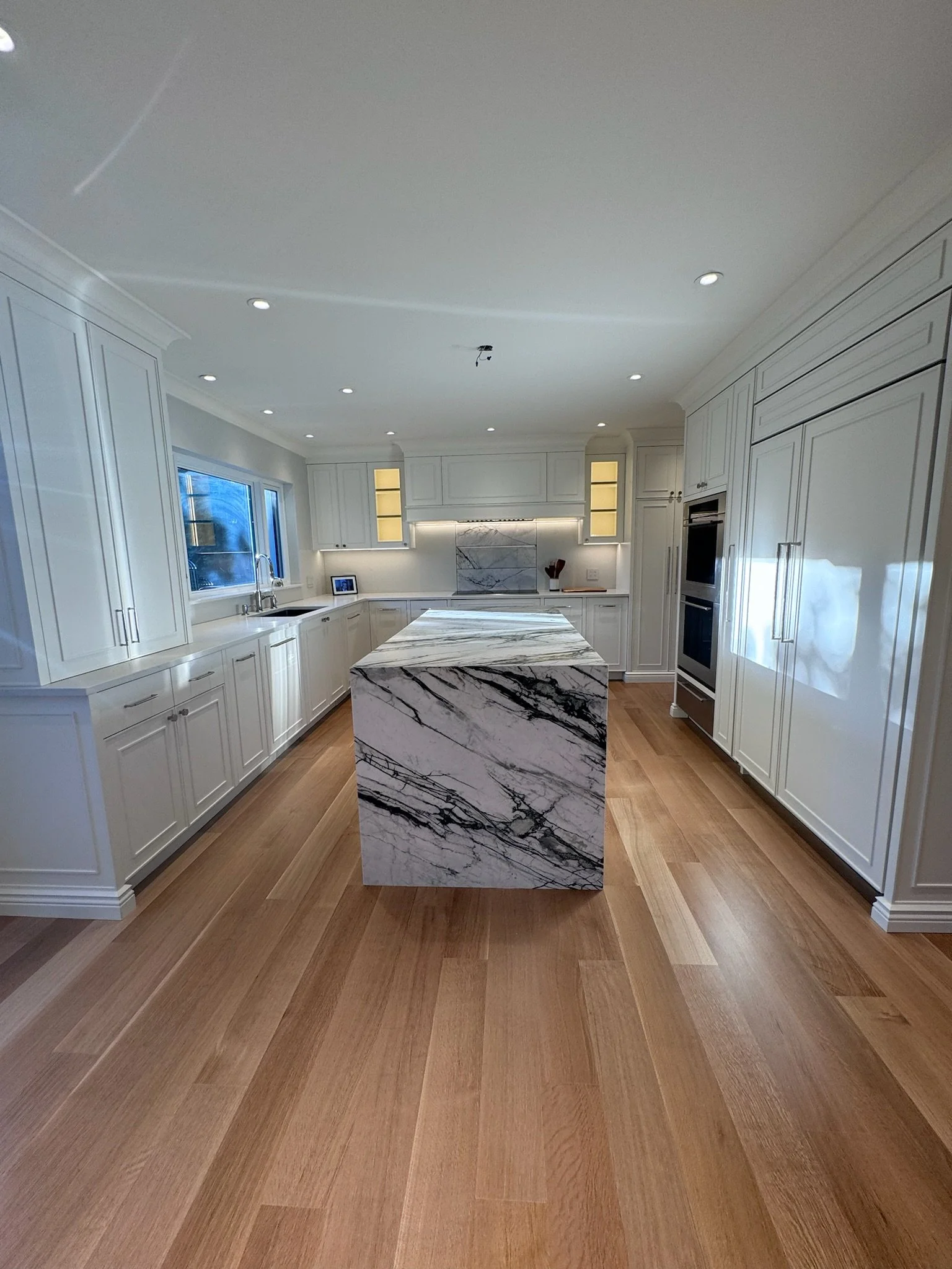 Modern kitchen with white cabinets, marble island, hardwood flooring, with built-in oven and microwave, and a small framed photo on the countertop.