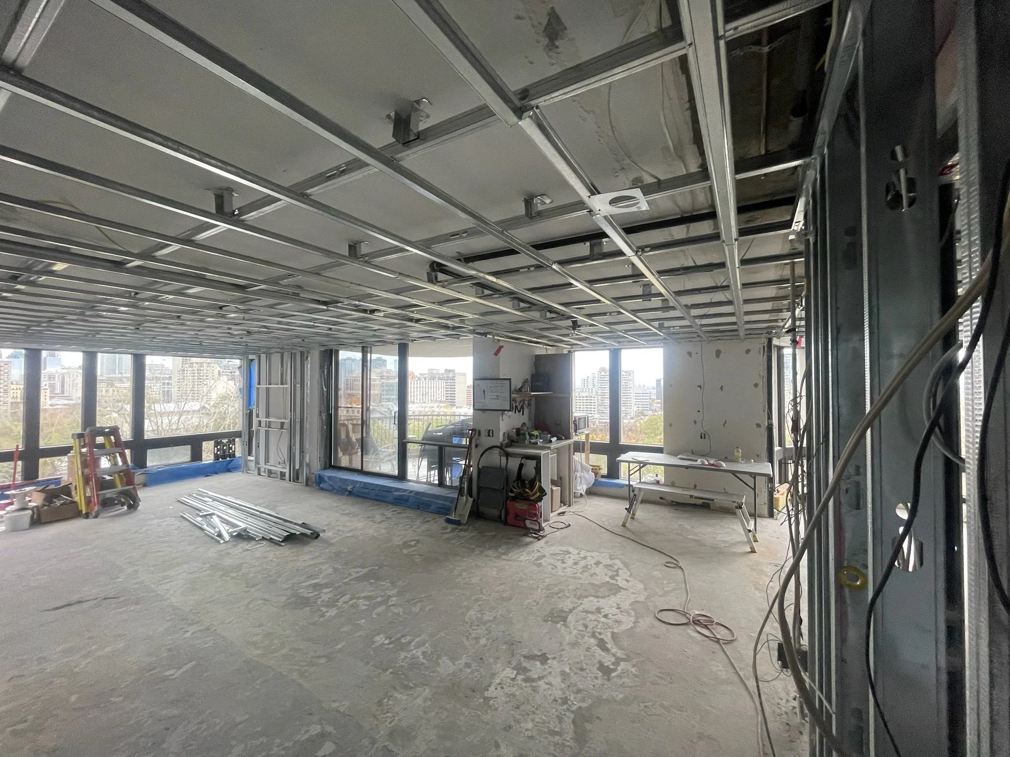 Interior view of a high-rise building under construction with exposed ceiling framework, construction materials, and tools scattered on the unfinished floor, large windows offering a cityscape view.
