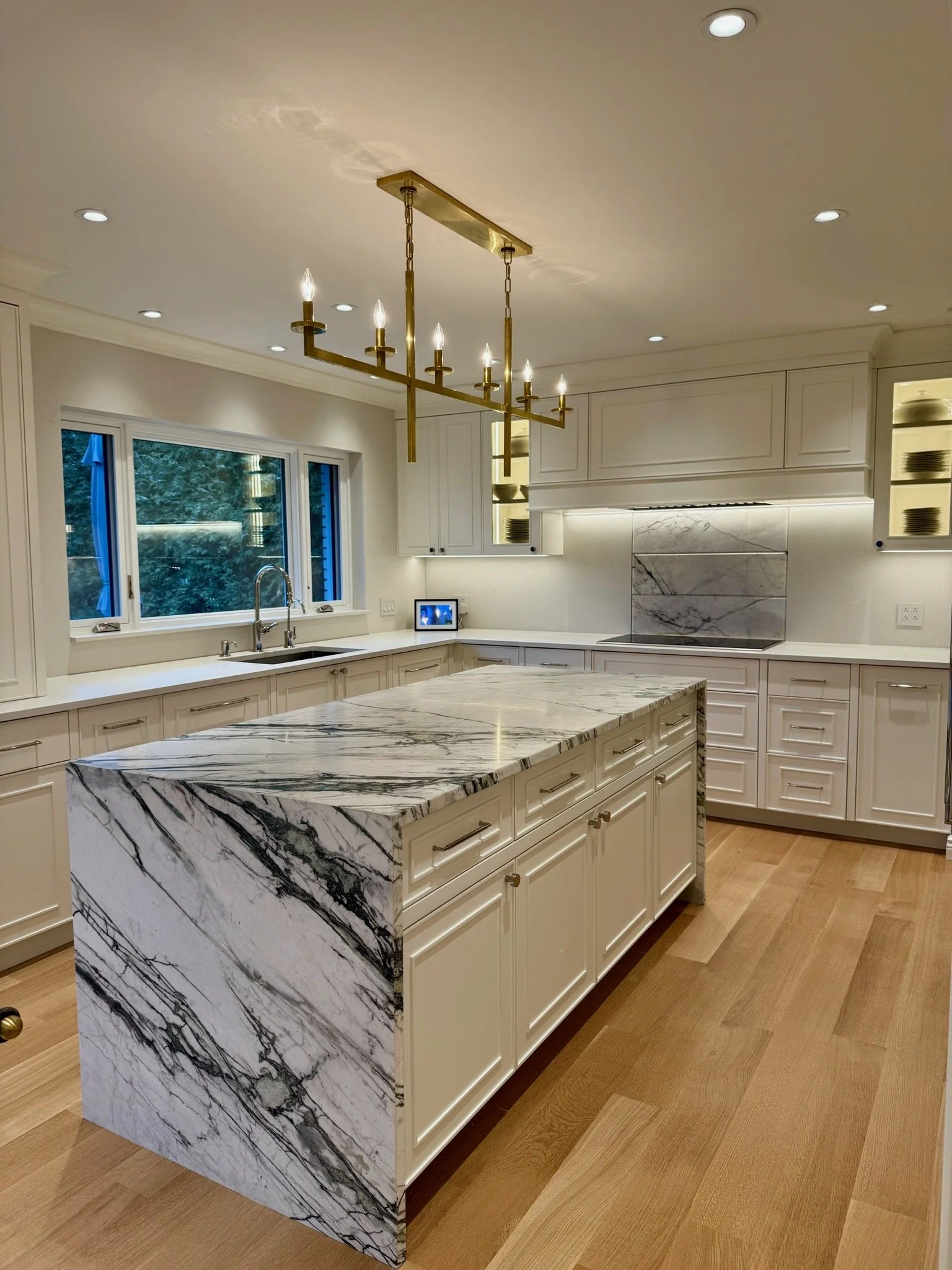 Modern kitchen with white cabinets, marble island, large window, and brass chandelier.