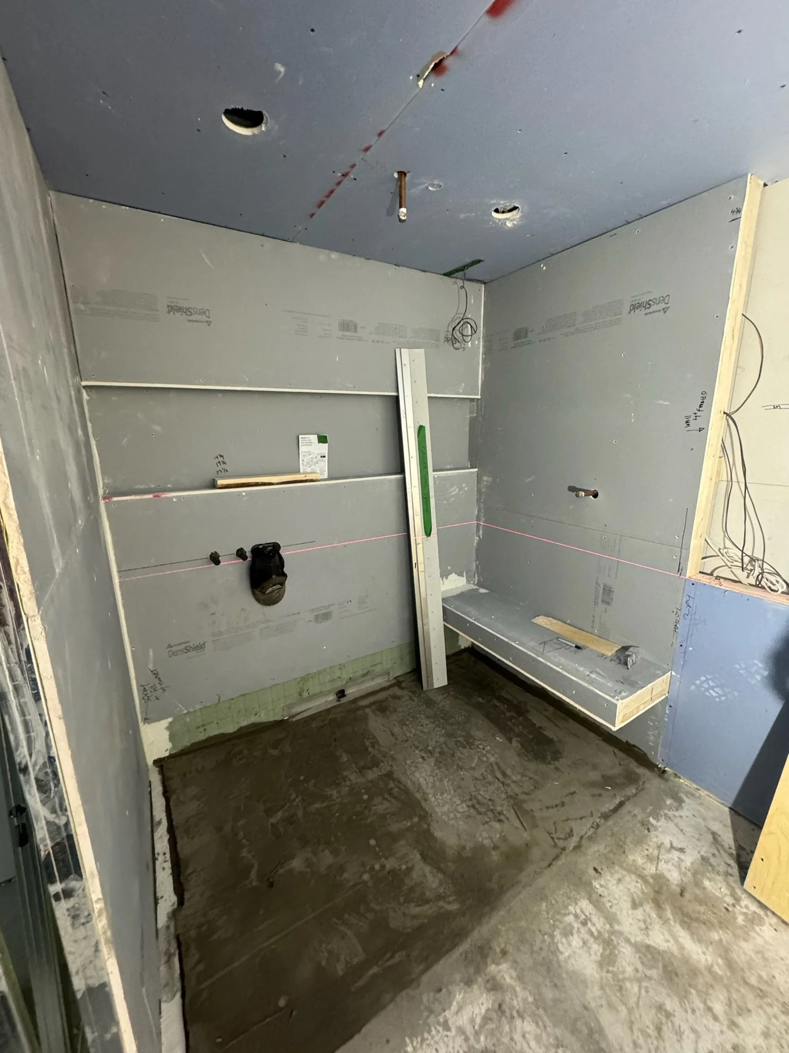Construction site showing an unfinished room with drywall installed on the walls and ceiling. The floor is rough concrete, and wires are hanging from the ceiling. There is a level leaning against the corner of the wall.