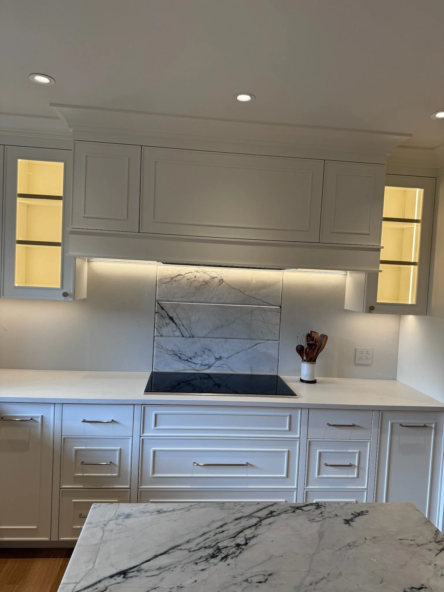 White kitchen with marble countertops, upper cabinets, built-in lighting, and a black stovetop.