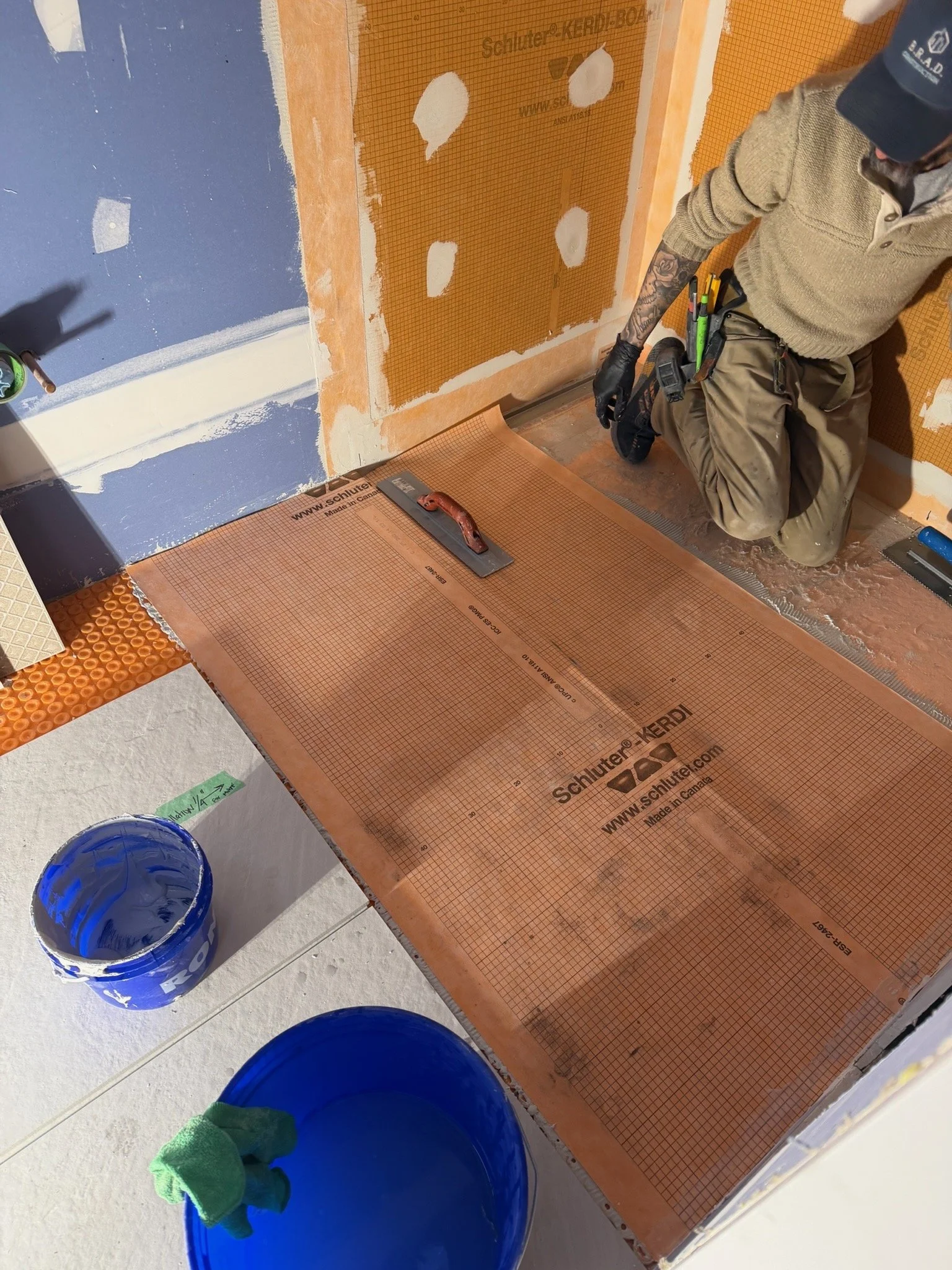 Person kneeling on floor applying a waterproof barrier sheet around a shower area, with buckets of blue sealant nearby.