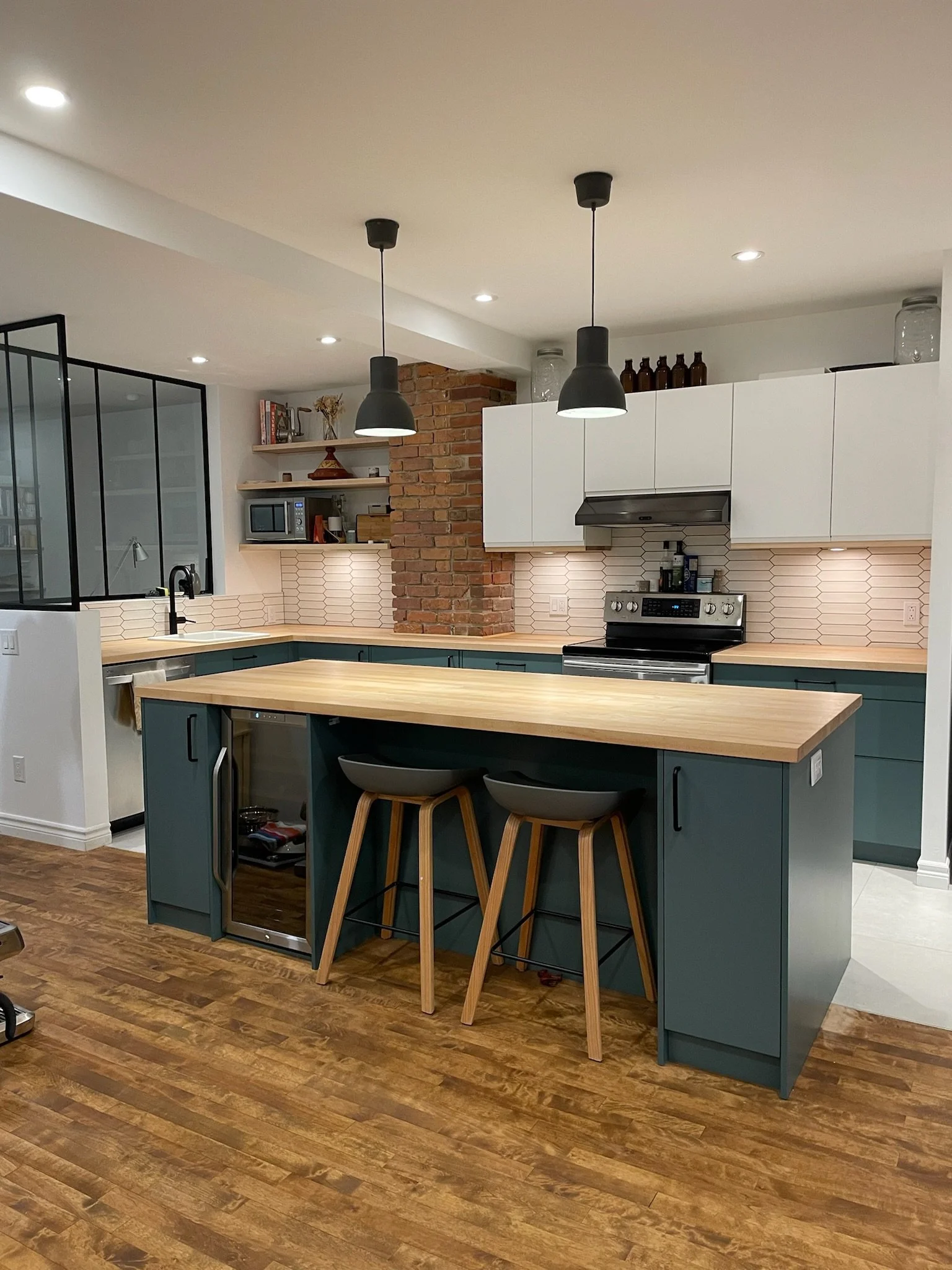 Modern kitchen with an island, blue cabinets, white upper cabinets, brick accent wall, open shelving, stainless steel range, black pendant lights, wood flooring, and bar stools.