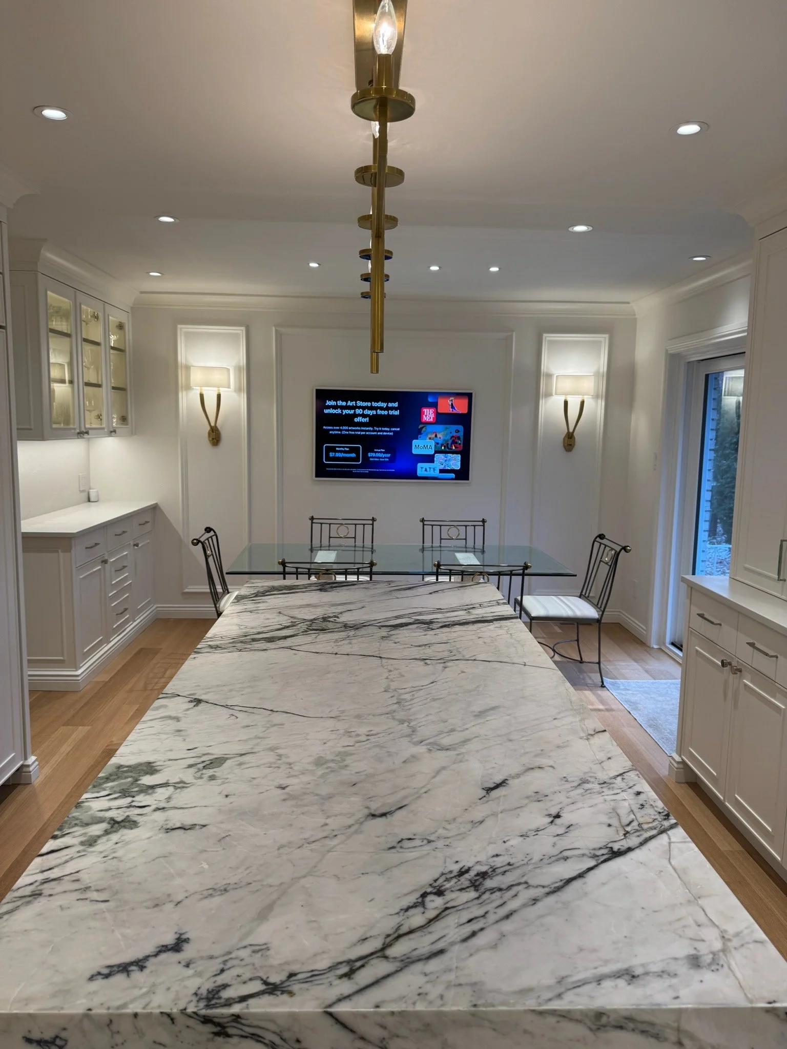 A kitchen dining area with a marble countertop in the foreground, white cabinets, a wall-mounted TV, and two wall sconces.