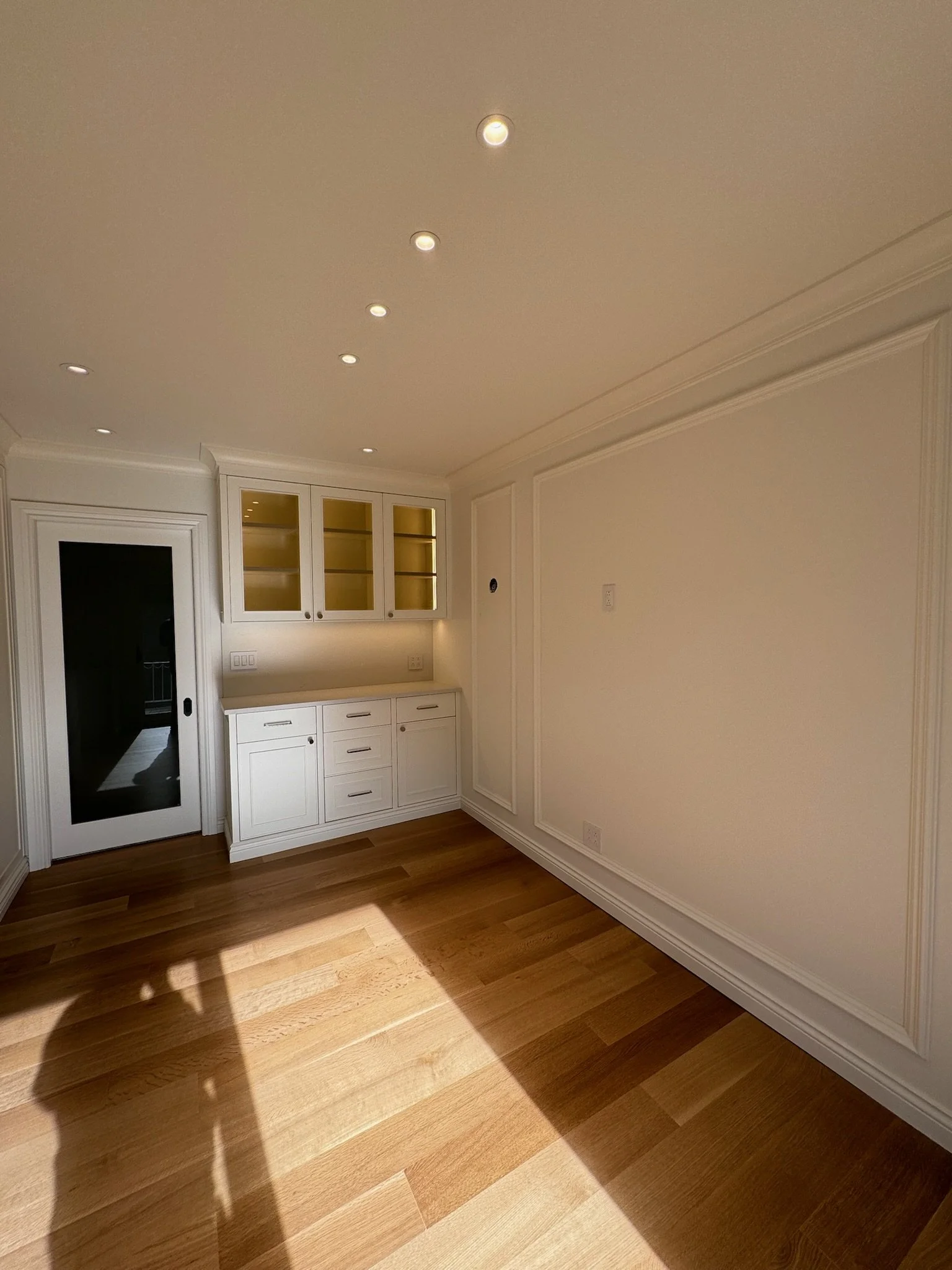 Empty room with white walls, wooden floor, built-in white cabinets with glass doors, and ceiling with multiple recessed lights.