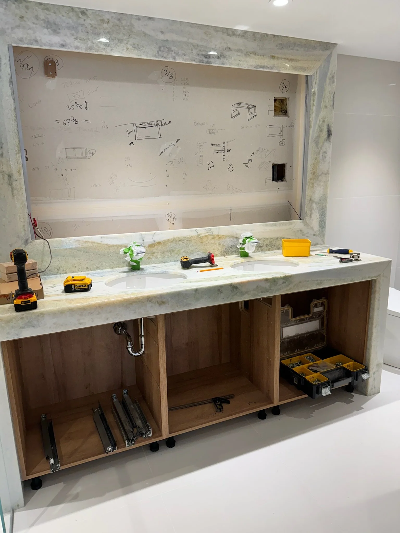 Bathroom vanity cabinet under construction with tools and supplies on and around it, wall behind with handwritten notes and sketches, open space for two sinks, electrical outlets, plumbing pipes visible underneath.