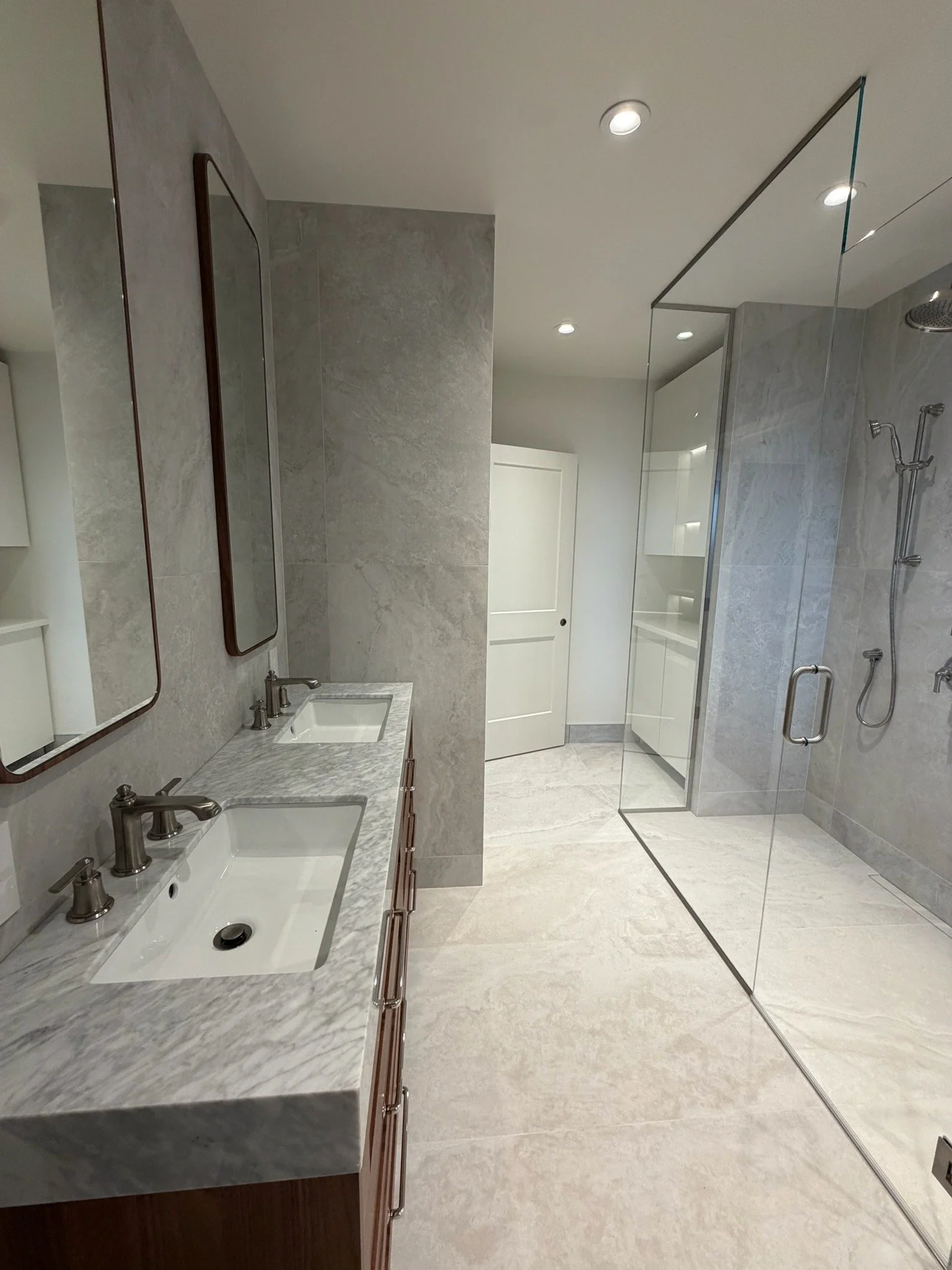 Modern bathroom with double vanity sinks with marble countertops, two mirrors, and a walk-in shower with glass doors, gray marble tiles, and multiple showerheads.