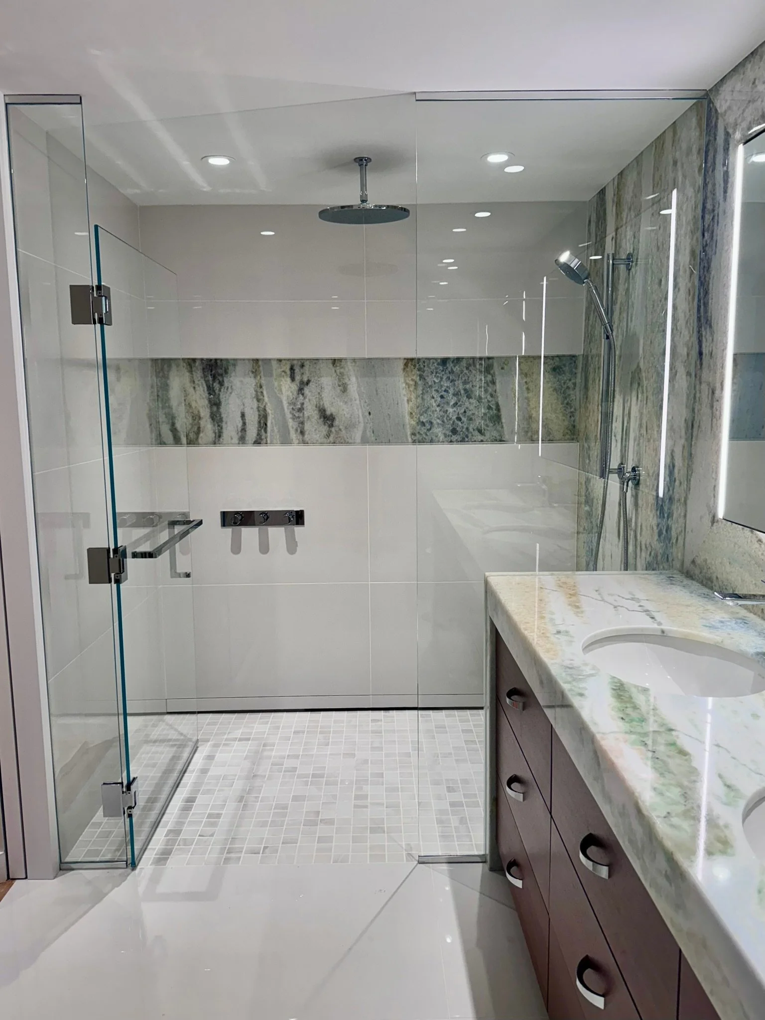 Modern bathroom with glass shower enclosure, marble wall tiles, white tile floor, and a marble countertop with a sink and vanity cabinet.