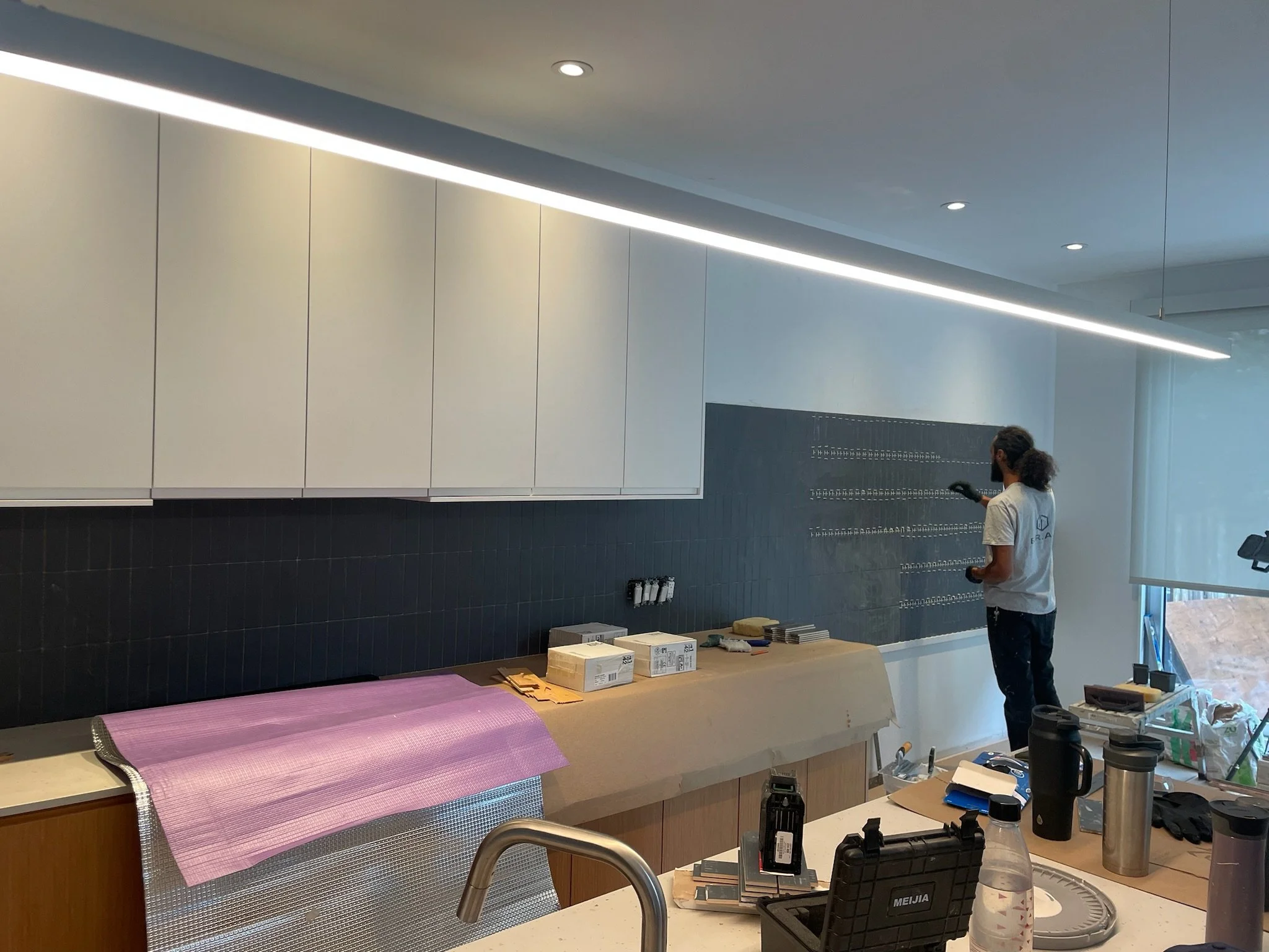 A person wearing a white shirt and black gloves is working in a modern, well-lit interior space, possibly under construction or renovation, with white cabinets, a black tiled wall, and various tools and materials on the work surface.