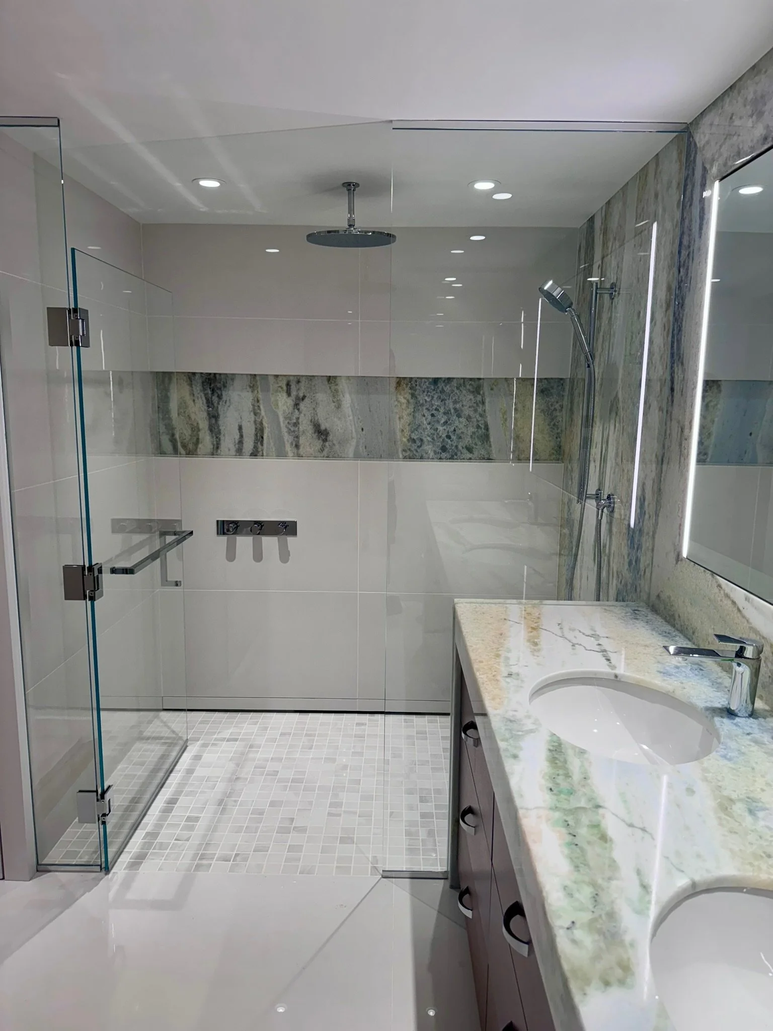 A modern bathroom with a glass-enclosed shower on the left, a showerhead hanging from the ceiling, a handheld shower on the right wall, a marble countertop with an oval sink and a mirror on the right wall, beige tiled walls, and a white tile floor.