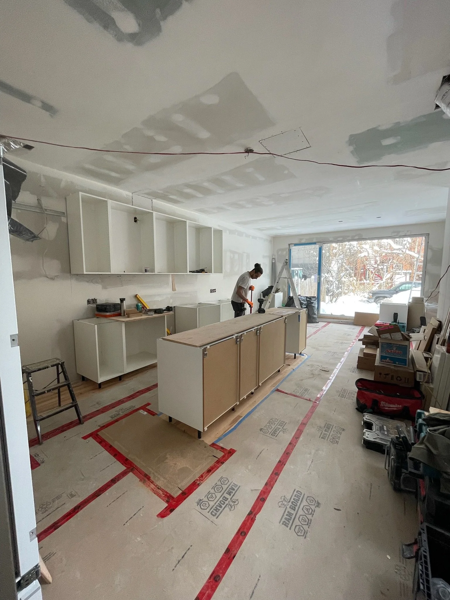 A person working on a kitchen renovation, installing cabinets and countertops, with construction materials and tools around, and a large window showing a snowy outdoor scene.