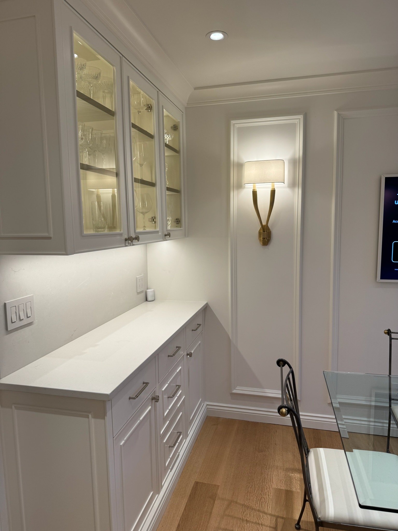 A corner of a white kitchen with white cabinets, a white countertop, a wall with decorative molding, a wall-mounted gold and white sconce light, a flat screen TV on the wall, a glass table with black chairs, and hardwood flooring.