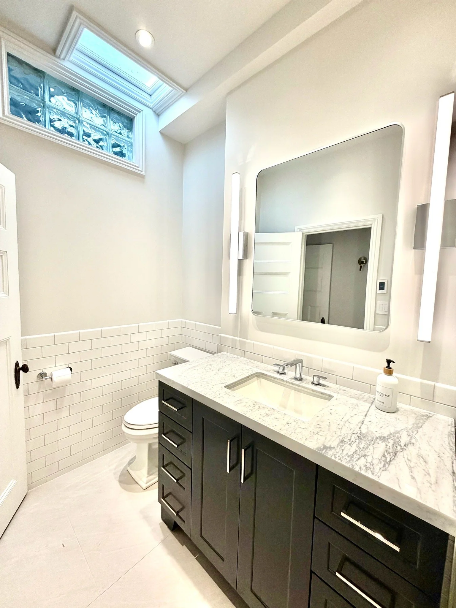 Modern bathroom with a large mirror, white subway tile on the walls, a vanity with black cabinets, a granite countertop, a sink, a soap dispenser, a toilet, a skylight, and a window.