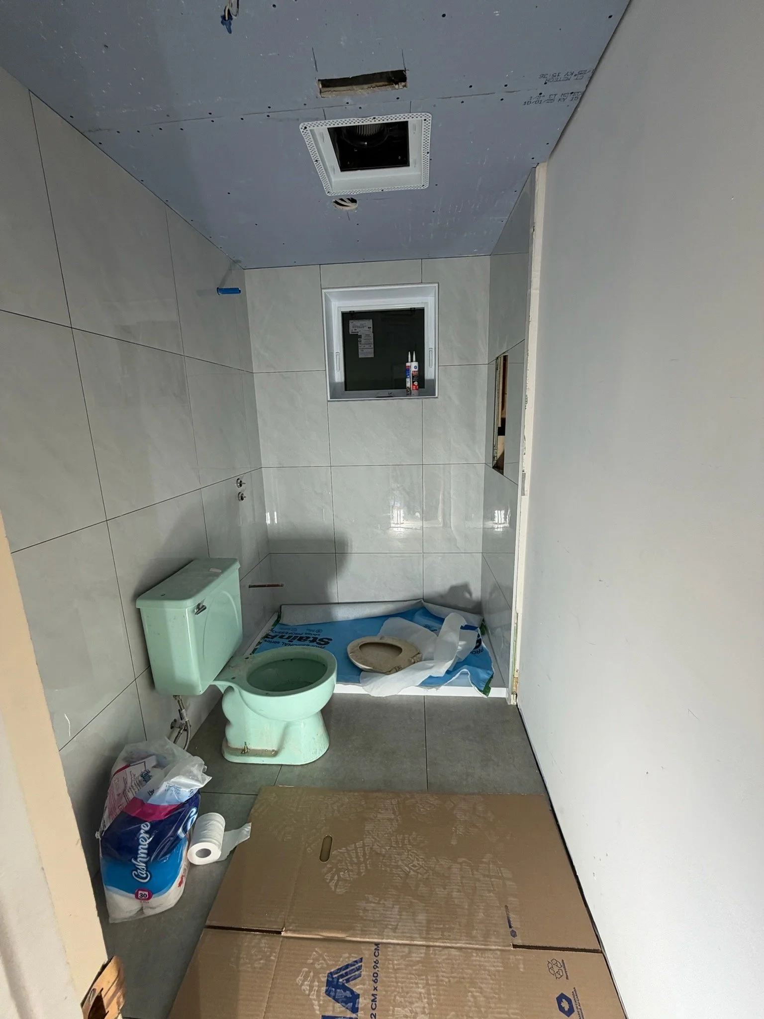 Bathroom under renovation with a mint green toilet, toilet paper roll, construction materials and tools, partially installed drywall, and a small window. The ceiling has a ceiling vent and an opening for a light fixture.